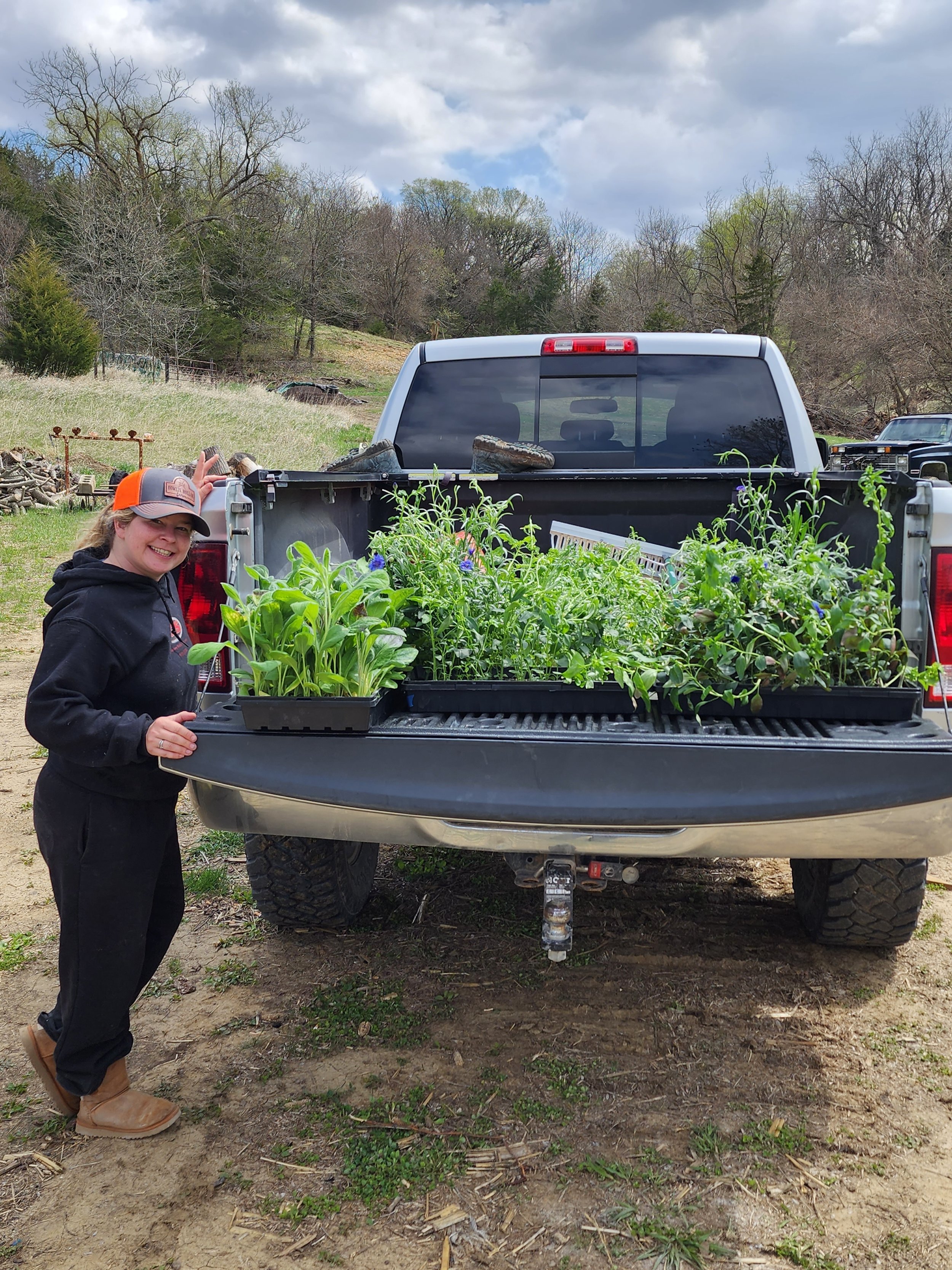 A woman standing beside a pickup truck with a bed full of potted plants and flowers outdoors on a cloudy day.