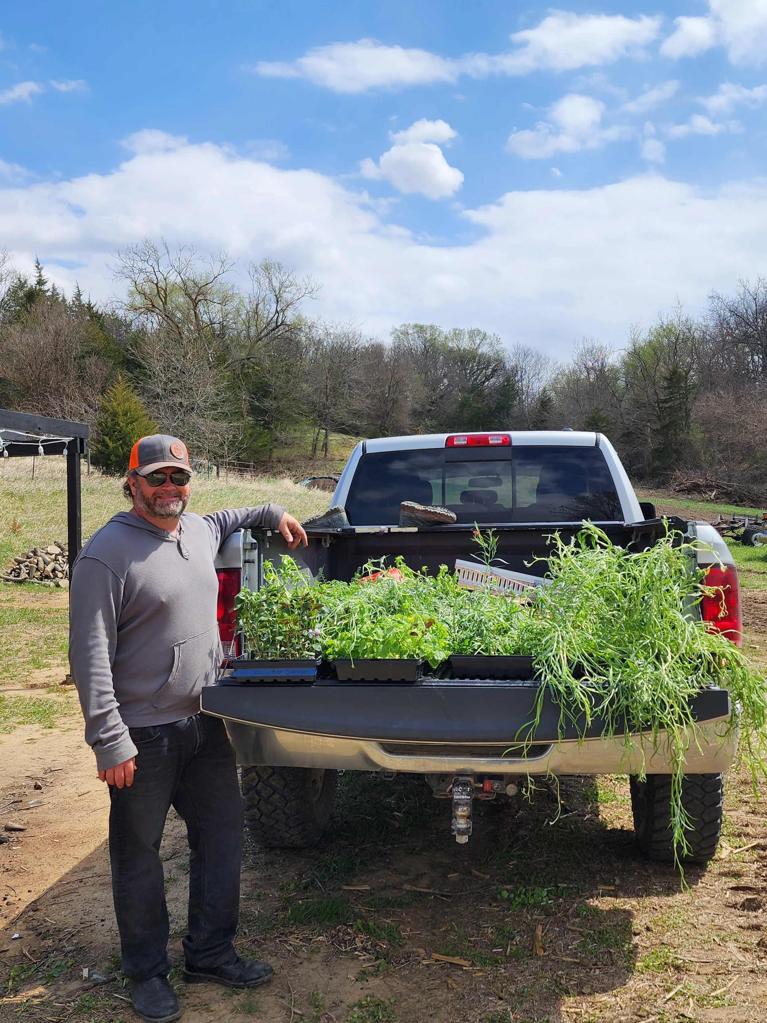 Man standing next to a pickup truck with a bed full of raised garden beds, outdoors on a partly cloudy day.