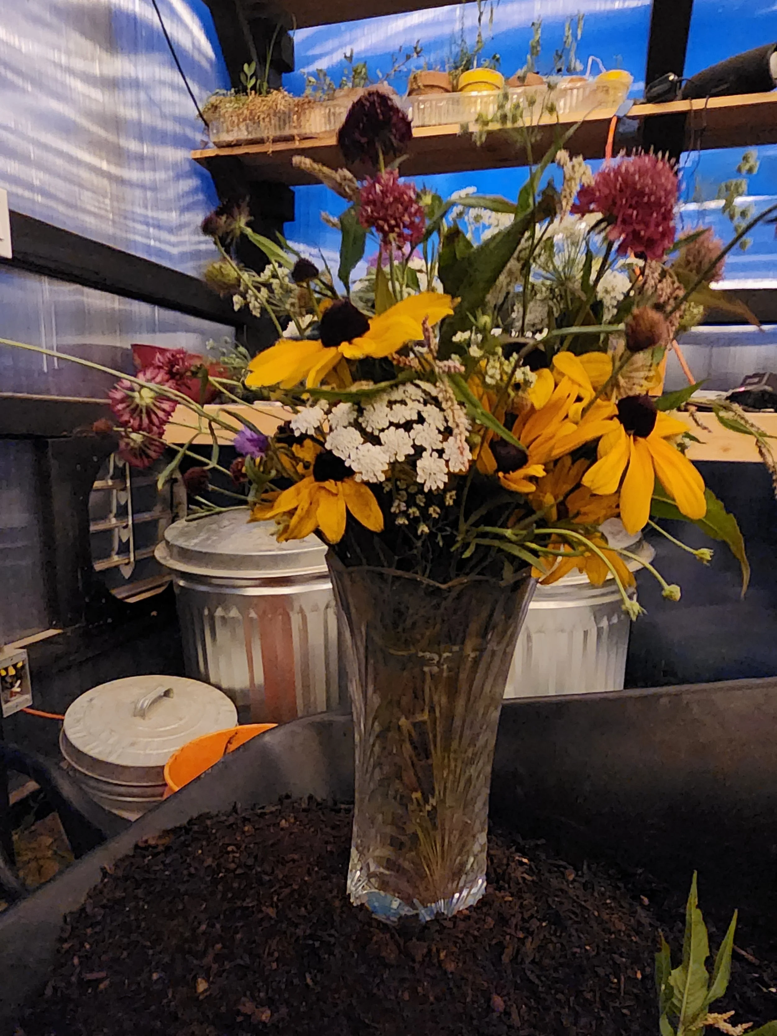 A tall, clear glass vase filled with a mixed bouquet of yellow, purple, pink, and white flowers on a dark surface.