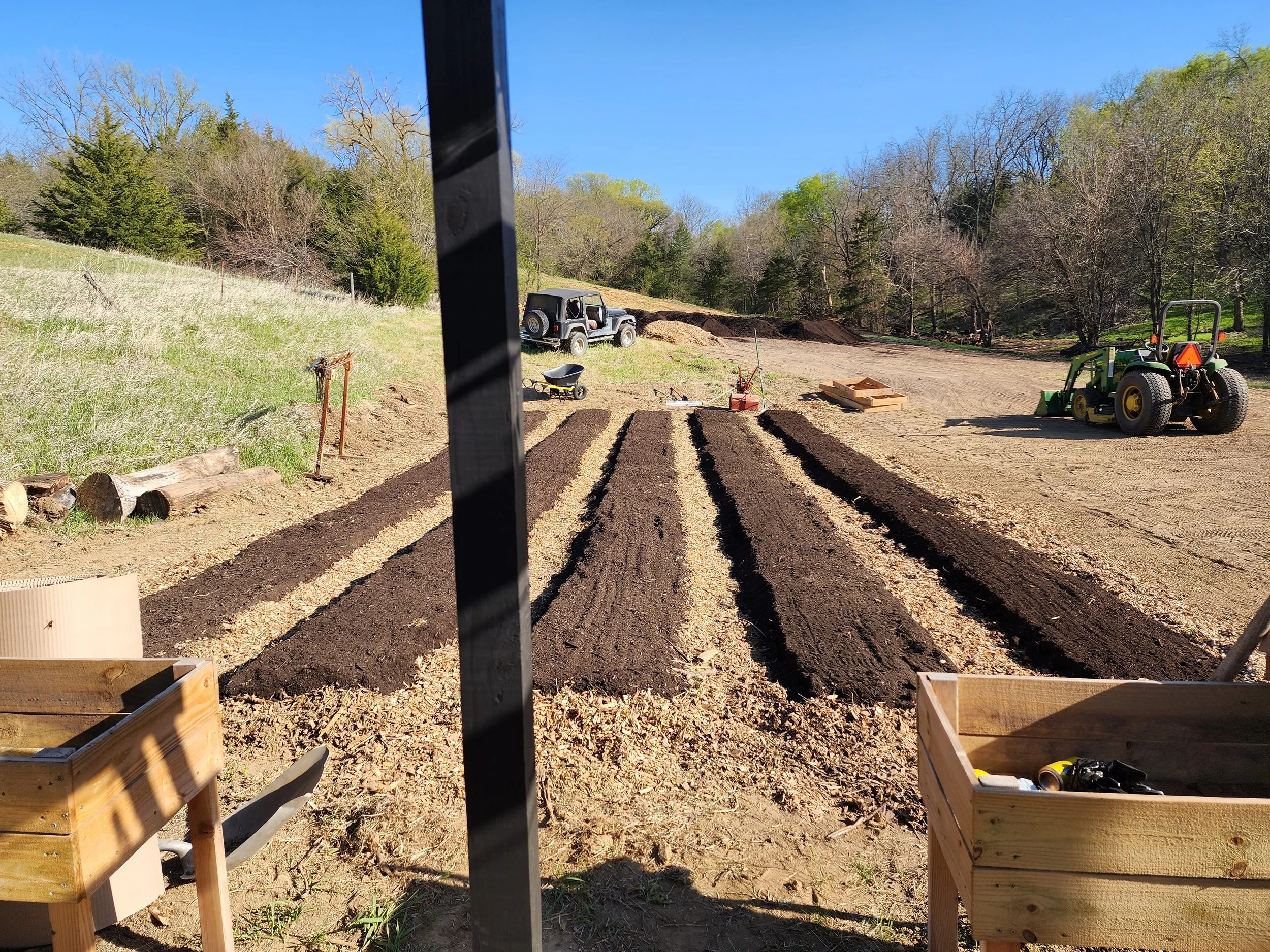 View of a garden bed being prepared with dark soil, with a tractor and other gardening tools nearby, and a wooded hillside in the background.