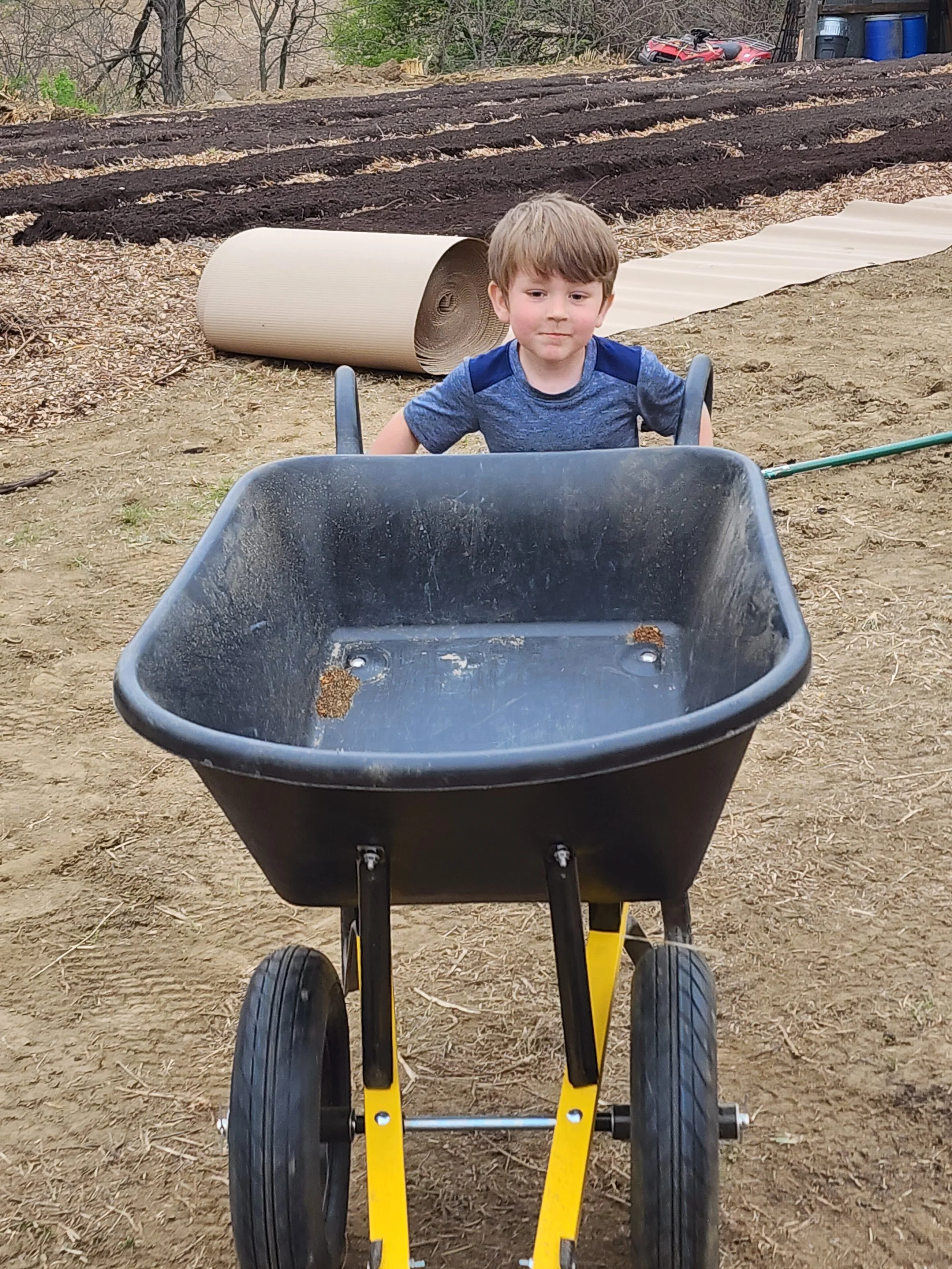 A young boy pushing an empty black wheelbarrow outdoors on dirt ground with a garden bed and fallen leaves in the background.