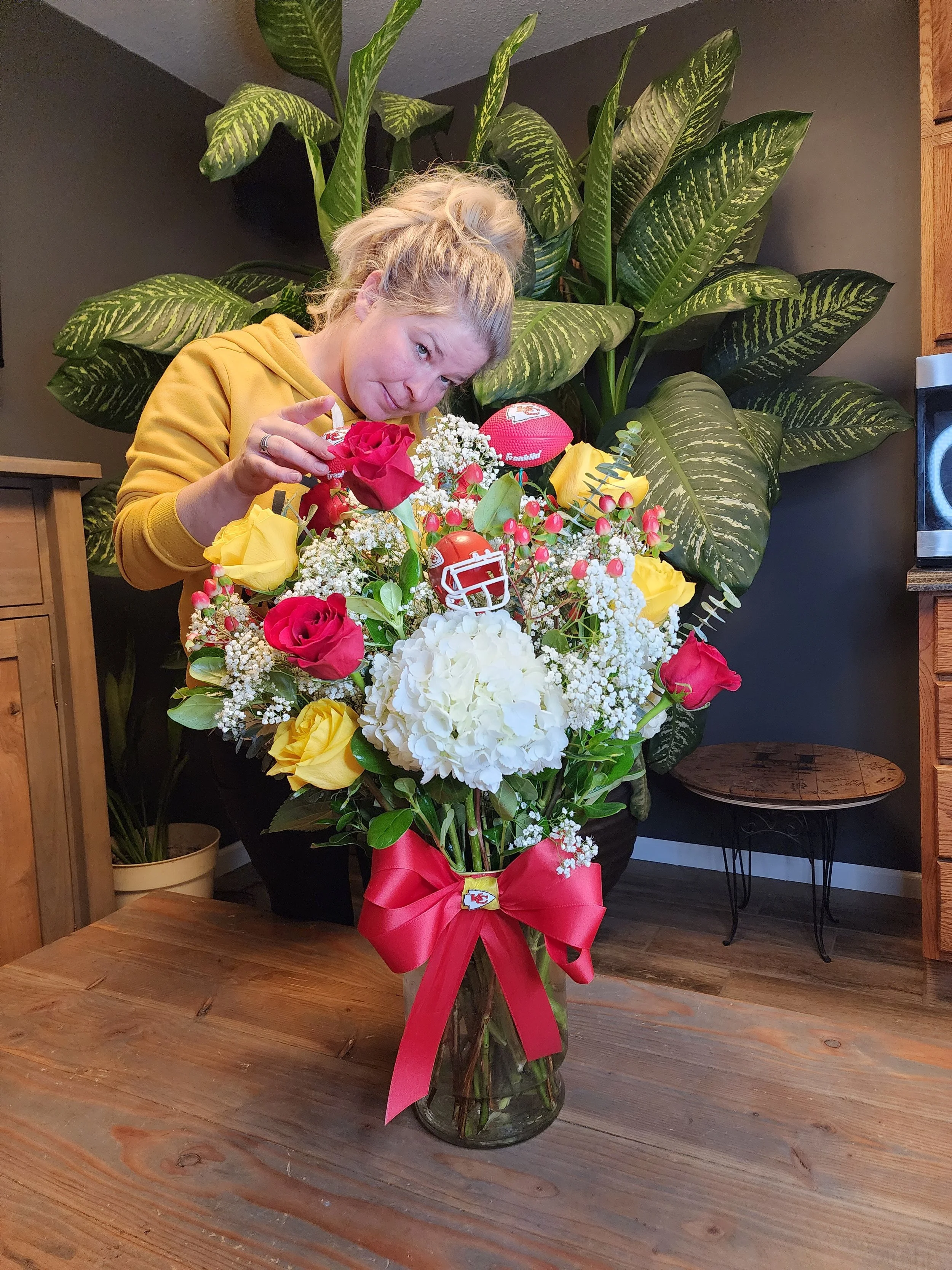 A woman in a yellow hoodie arranging a floral bouquet with pink roses, yellow roses, white hydrangeas, baby's breath, and small red berries, decorated with Kansas City Chiefs football-themed ornaments and a large red ribbon, on a wooden table.