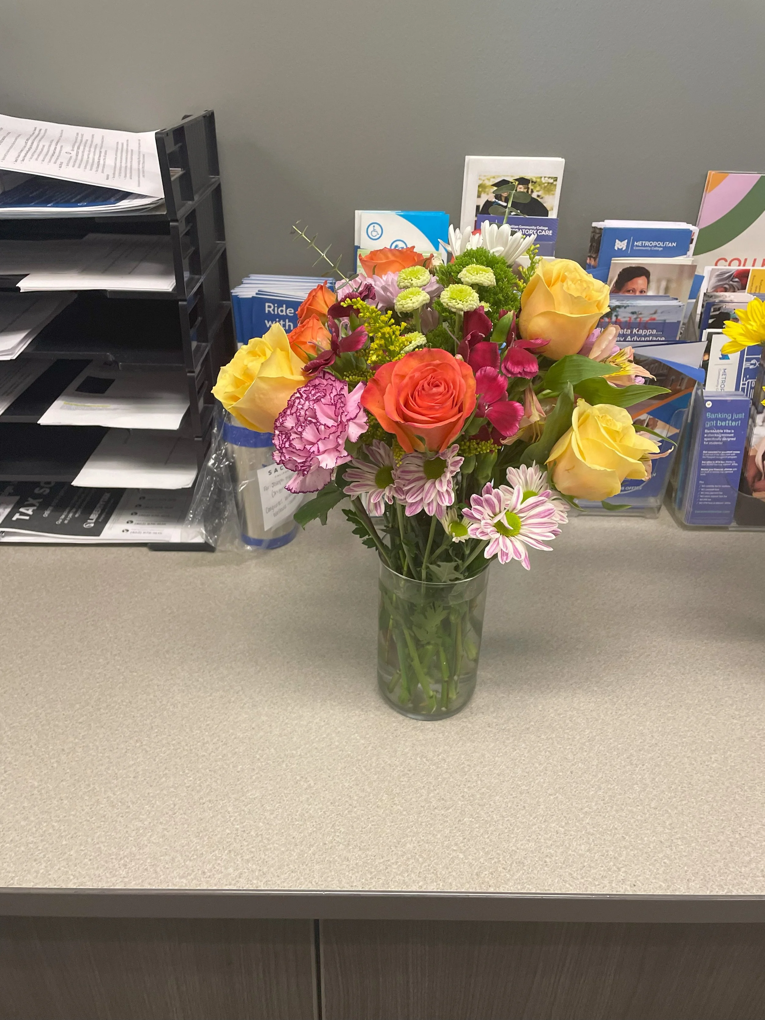 A colorful bouquet of flowers in a clear glass vase on a beige office desk, with brochures and papers in the background.