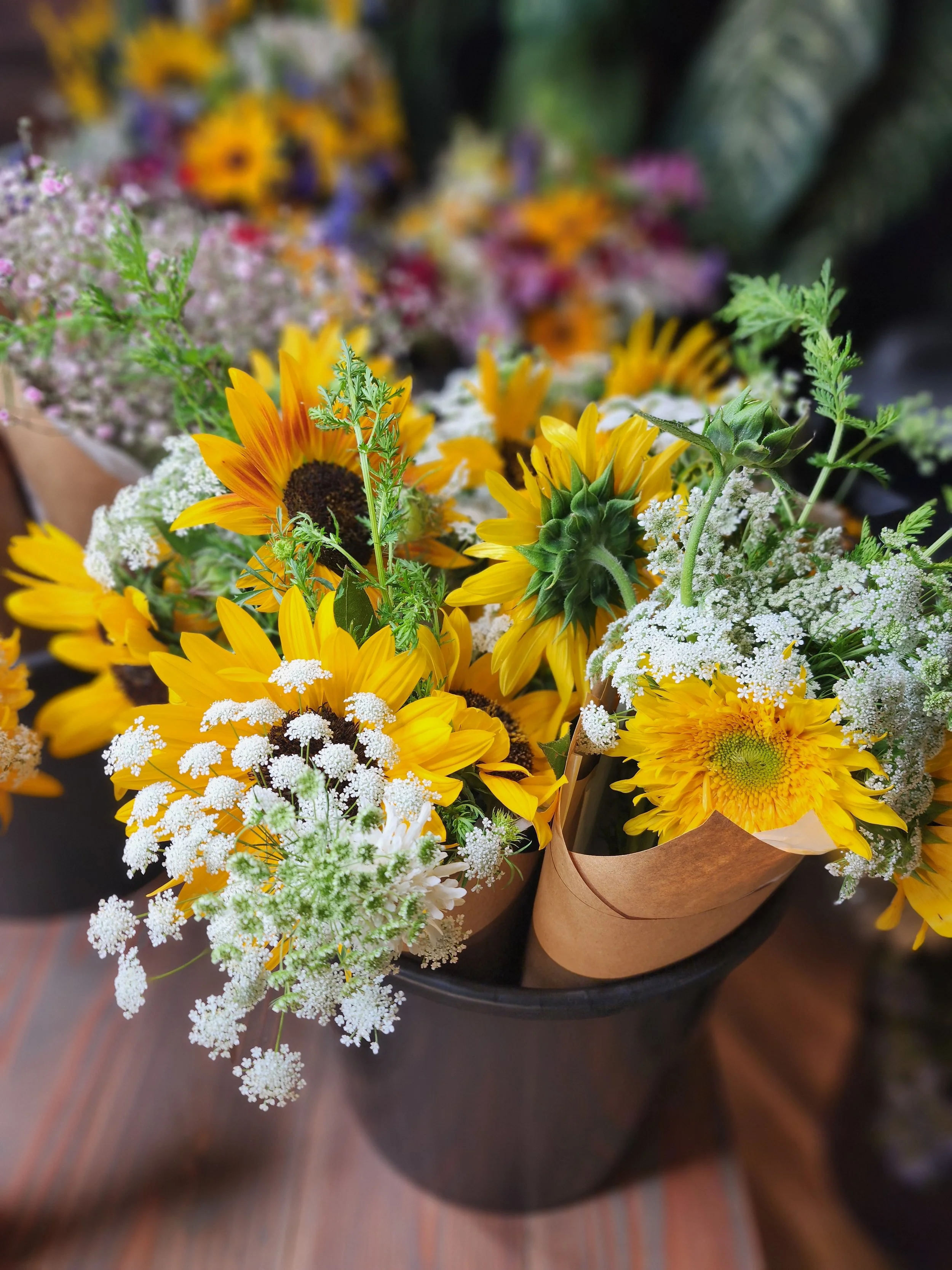 Bouquet of sunflowers, white flowers, and greenery in a black vase with light brown paper wrapping.