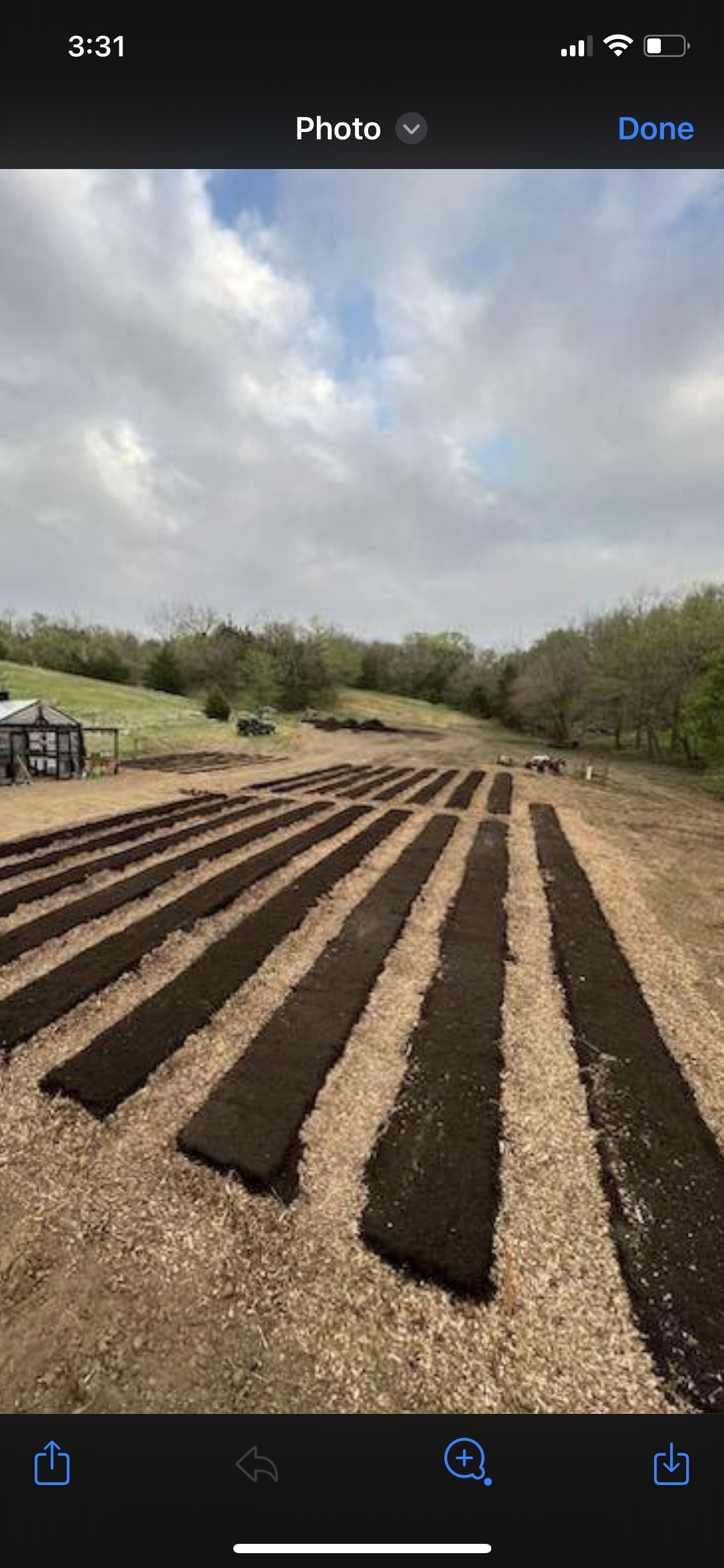 A farm field with freshly tilled dark soil arranged in evenly spaced long rows, surrounded by green trees and a cloudy sky.