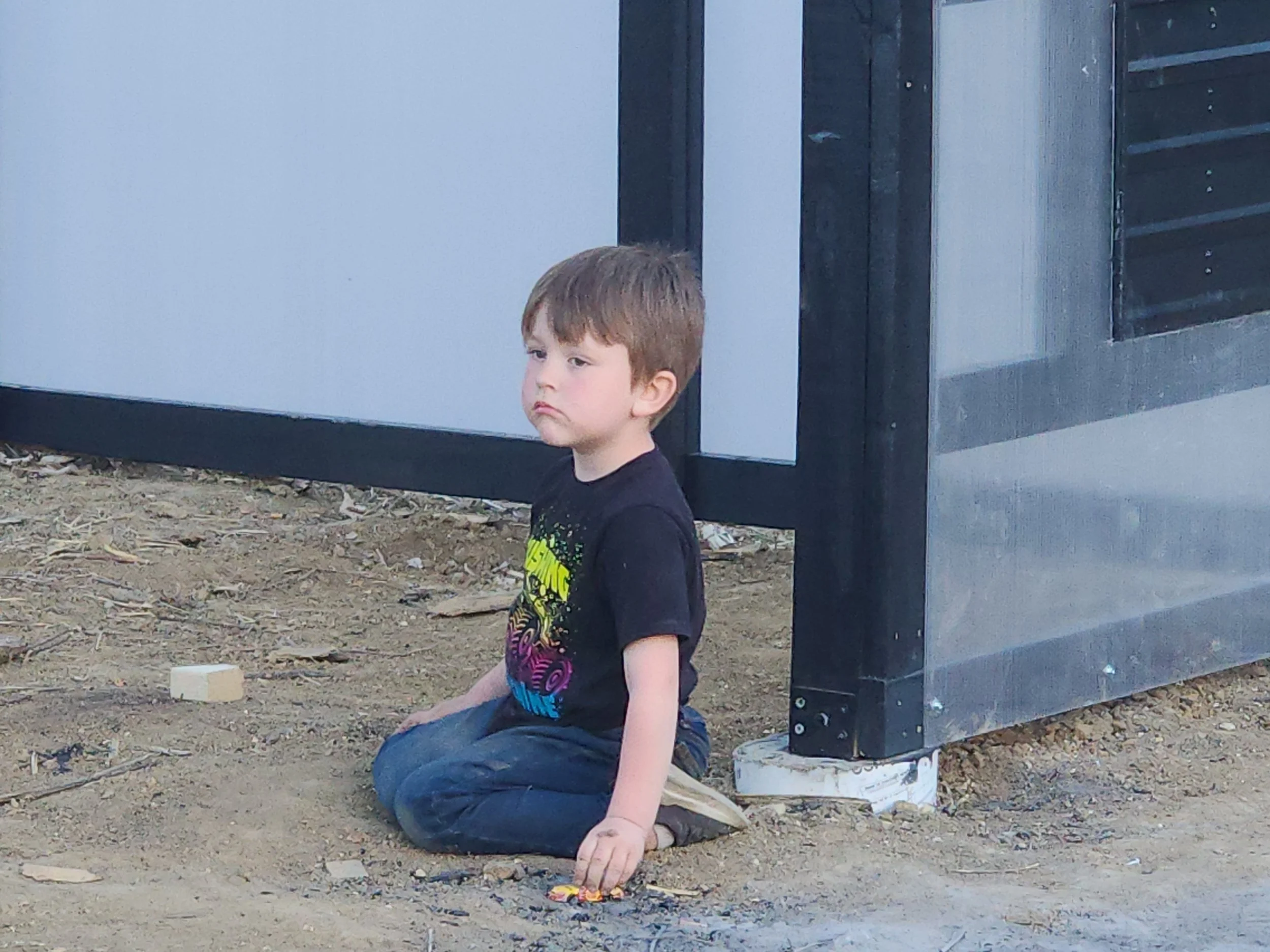 A young boy sitting on the ground outdoors near a modern black and white fence with a somber expression.