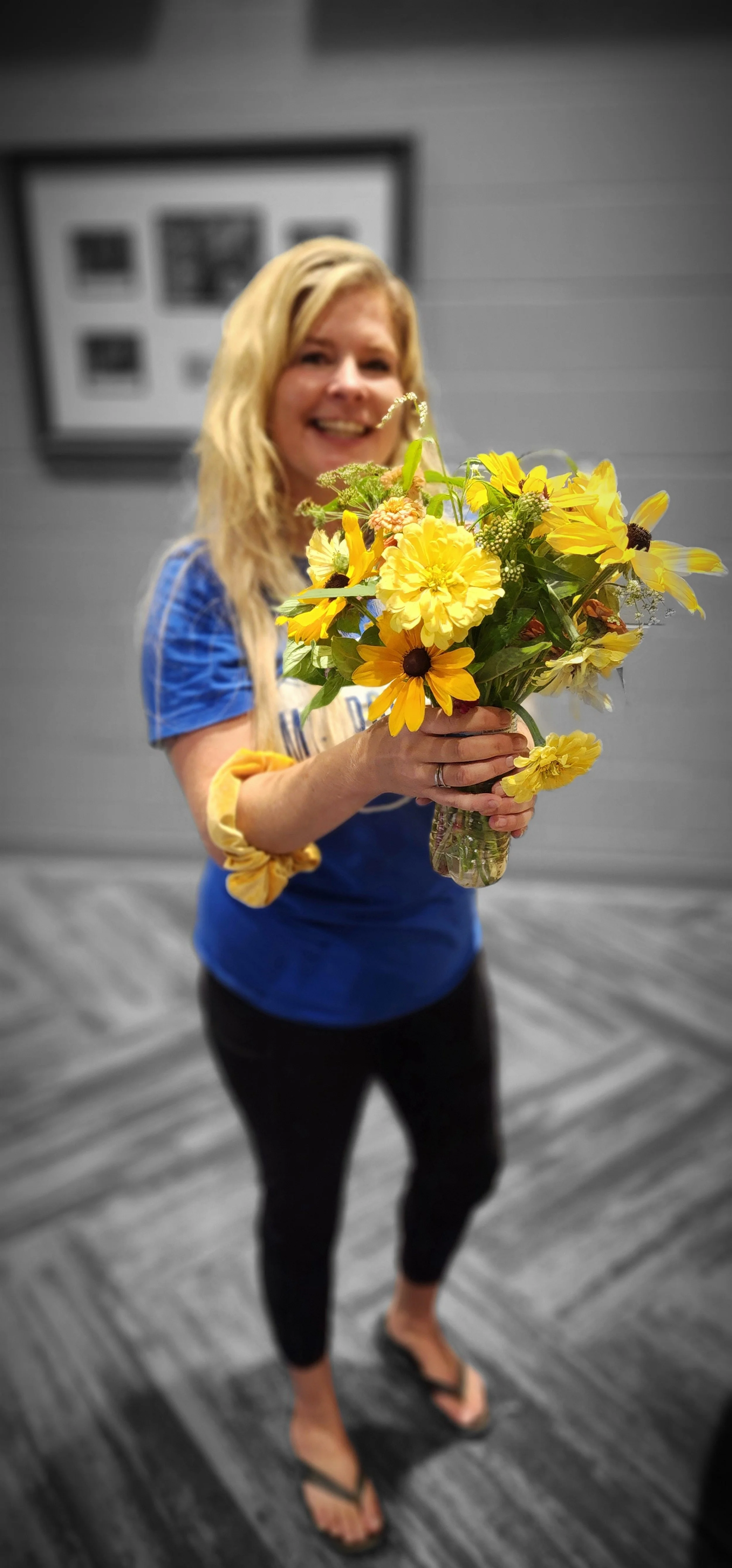 A woman with blonde hair wearing a blue shirt and black pants standing on a gray wooden floor, holding a bouquet of yellow flowers. The background is in black and white.