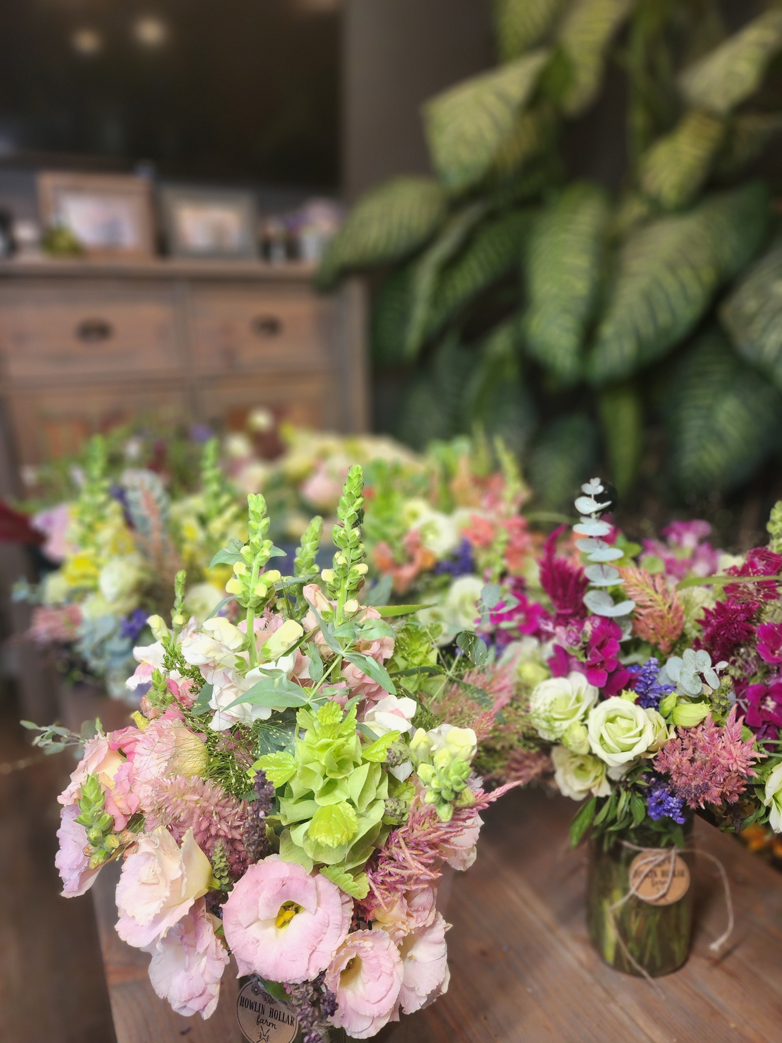 Colorful flower arrangements in glass jars on a wooden table with a blurred background of a wooden dresser and a large leafy plant