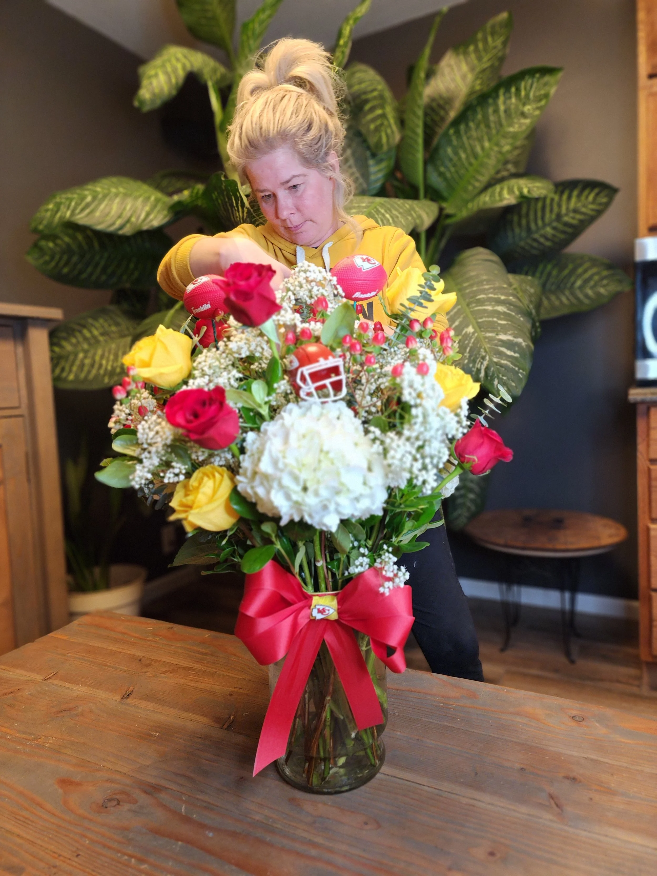 Woman in yellow hoodie arranging a sports-themed flower bouquet with football decorations and roses in a glass vase, on a wooden table.