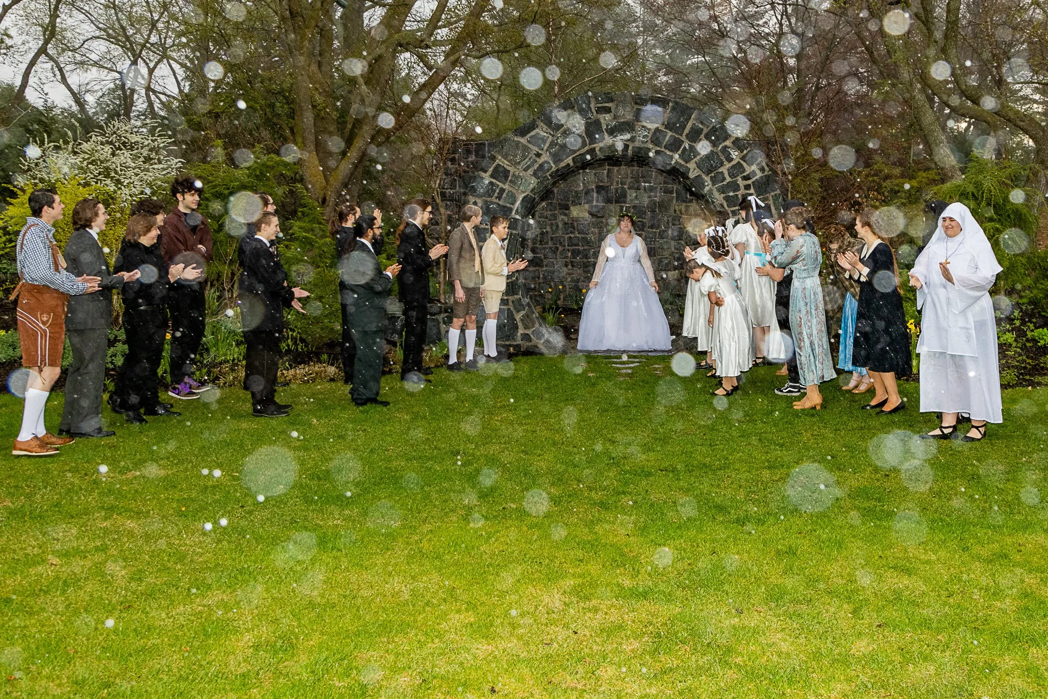 An actor dressed in a wedding dress is admired by the ensemble