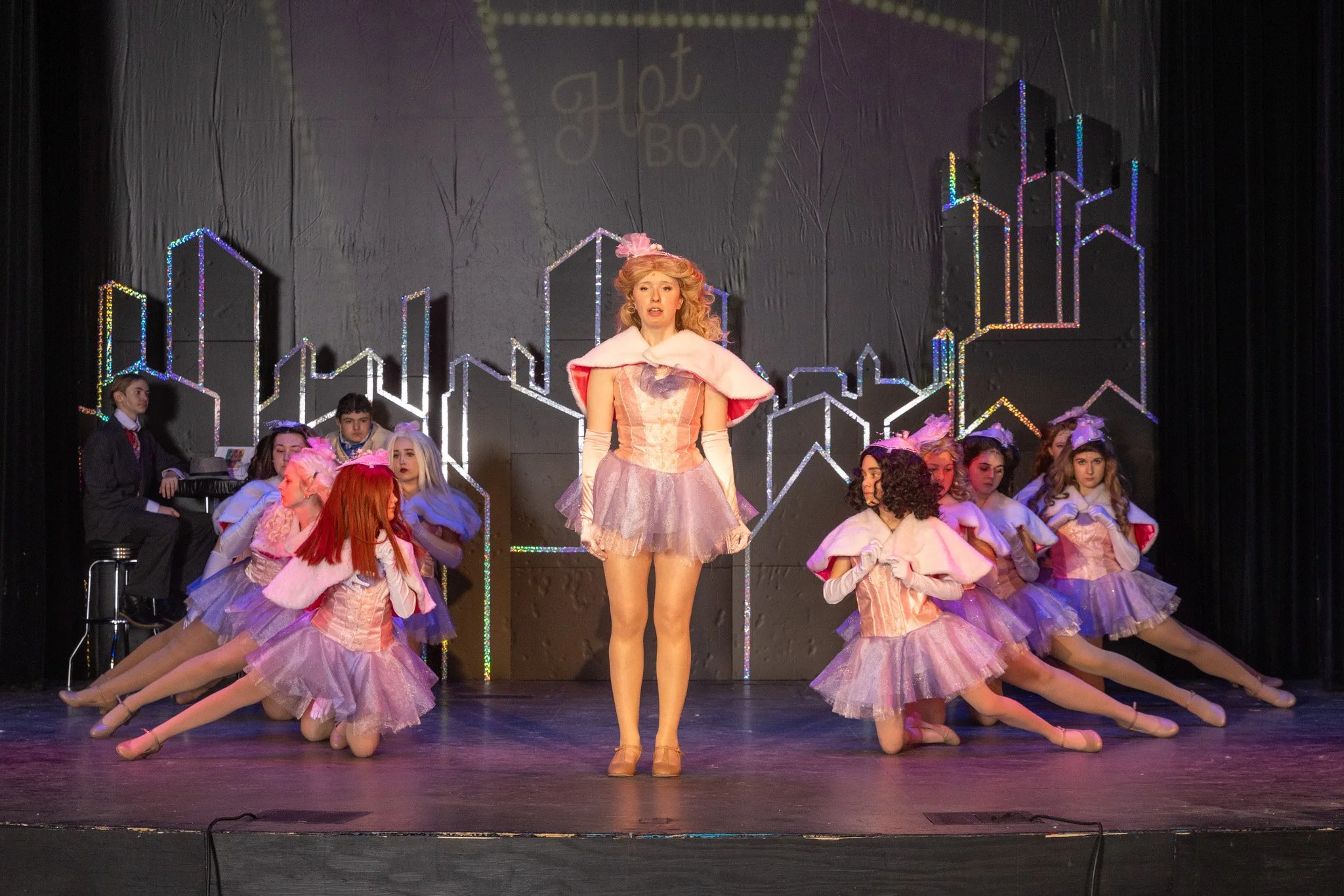An actor wearing a pink tutu and white fur wrap sings in the center of the stage as ensemble dancers in matching tutus pose on the ground behind her