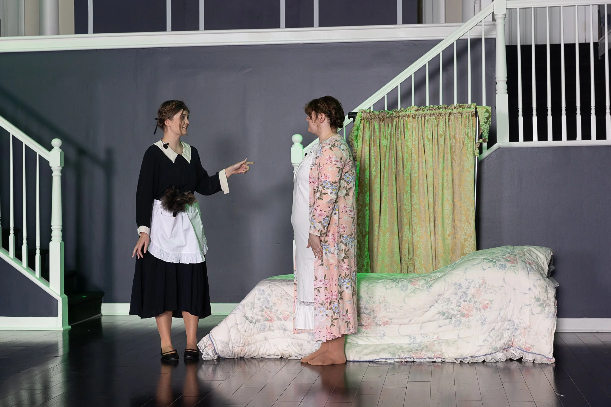 Two actors stand on stage and talk in front of a bed