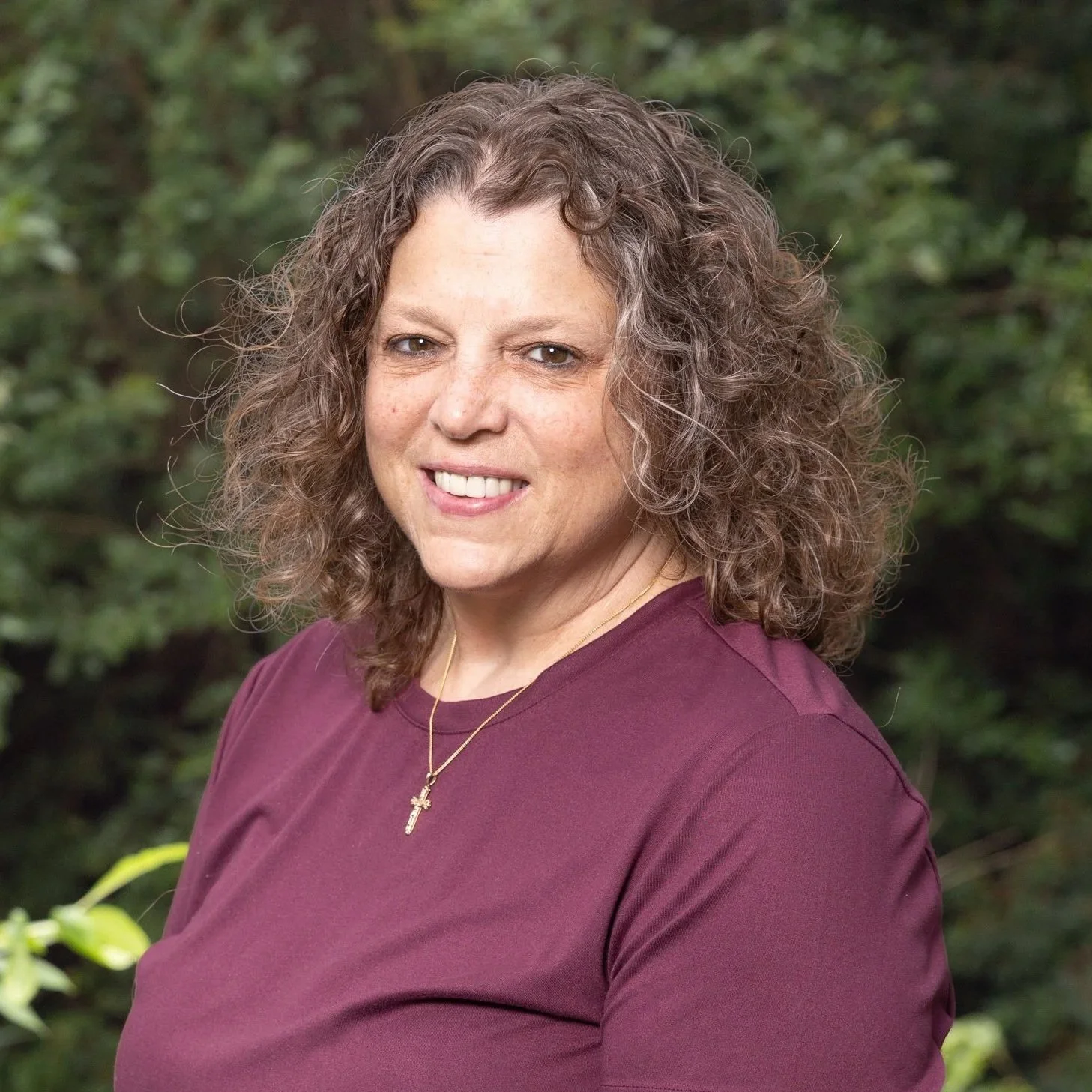 A woman with curly brown hair smiling outdoors, wearing a maroon shirt and a gold necklace with a cross pendant.