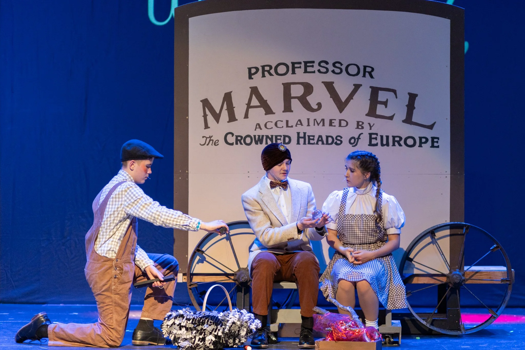 Three actors sit on a covered wagon in front of a fake fire
