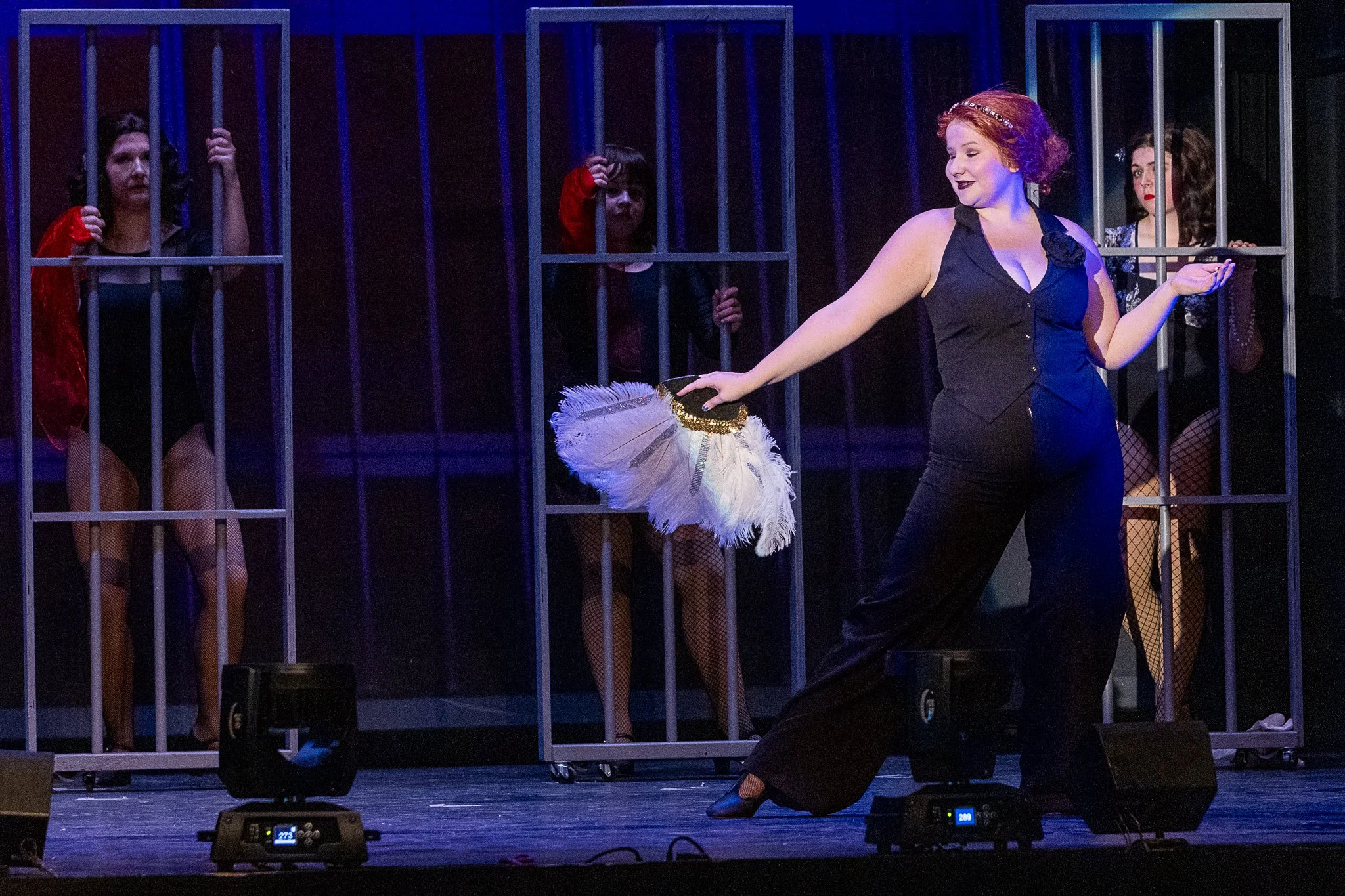 An actor dances with a large feather fan in front of three girls in jail cells