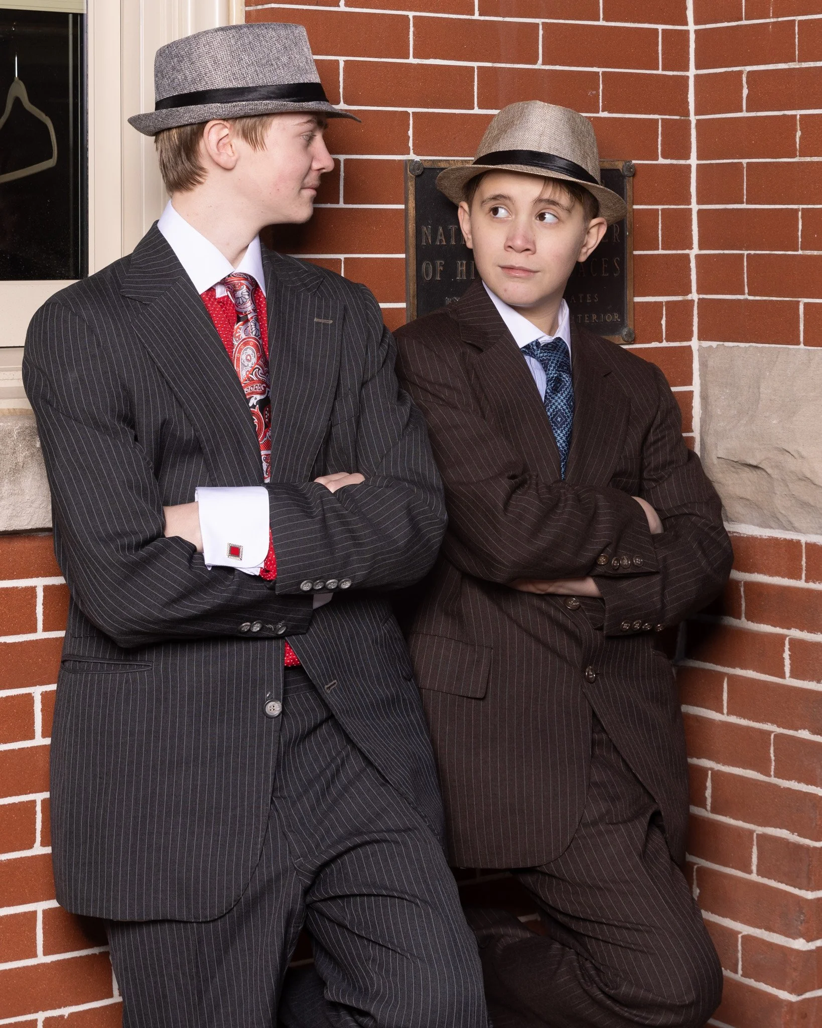 Two actors lean against a brick building and look at each other