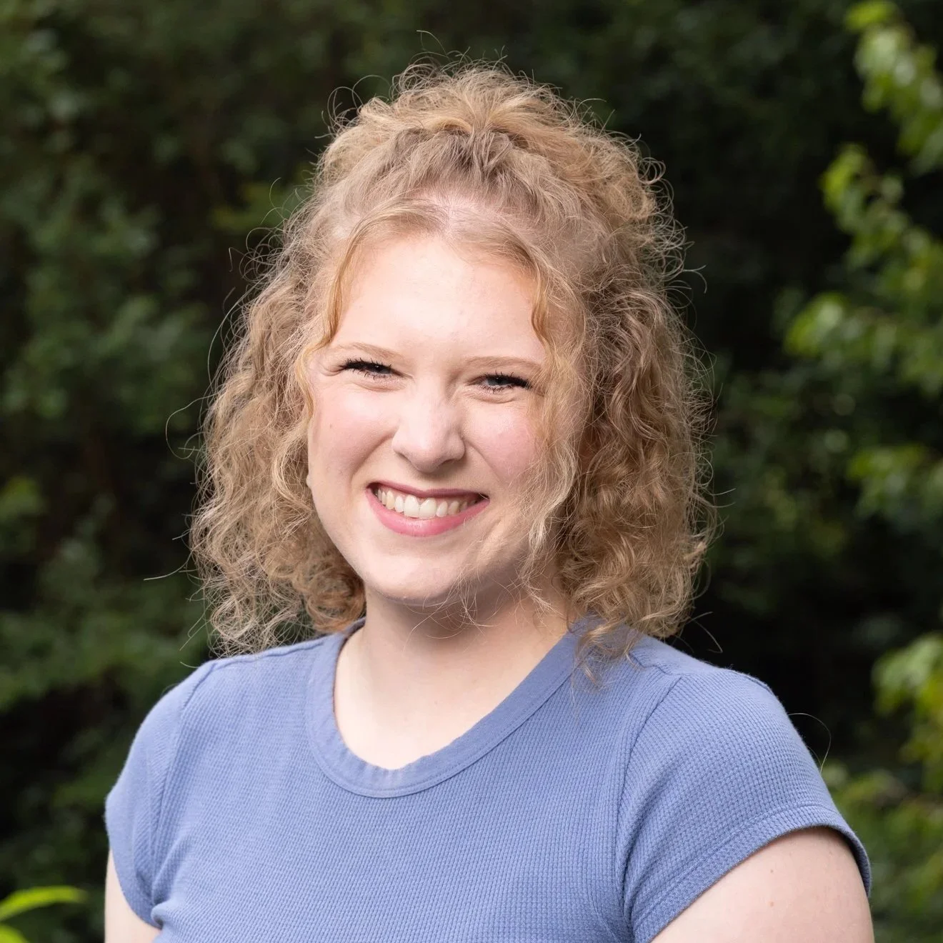 A young woman with curly red hair smiling outdoors against a green, leafy background.
