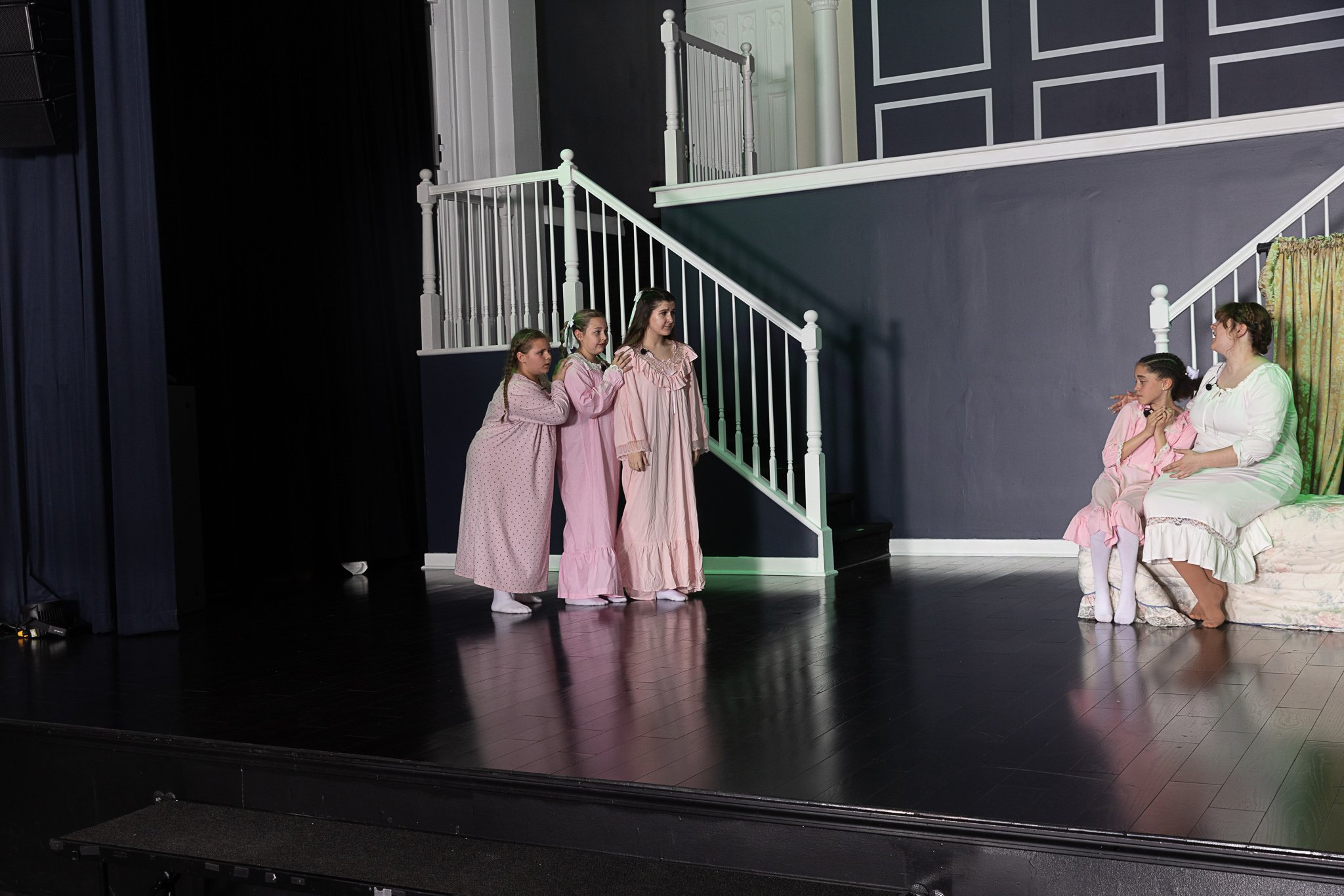 Two actors sit on a bed as three others talk to them from the other side of the stage