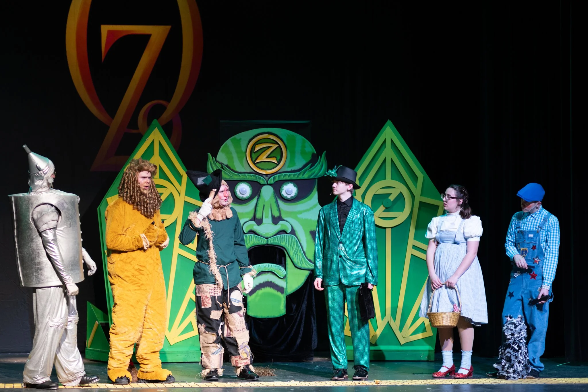 Six actors stand on a stage in front of a giant talking head and two green walls