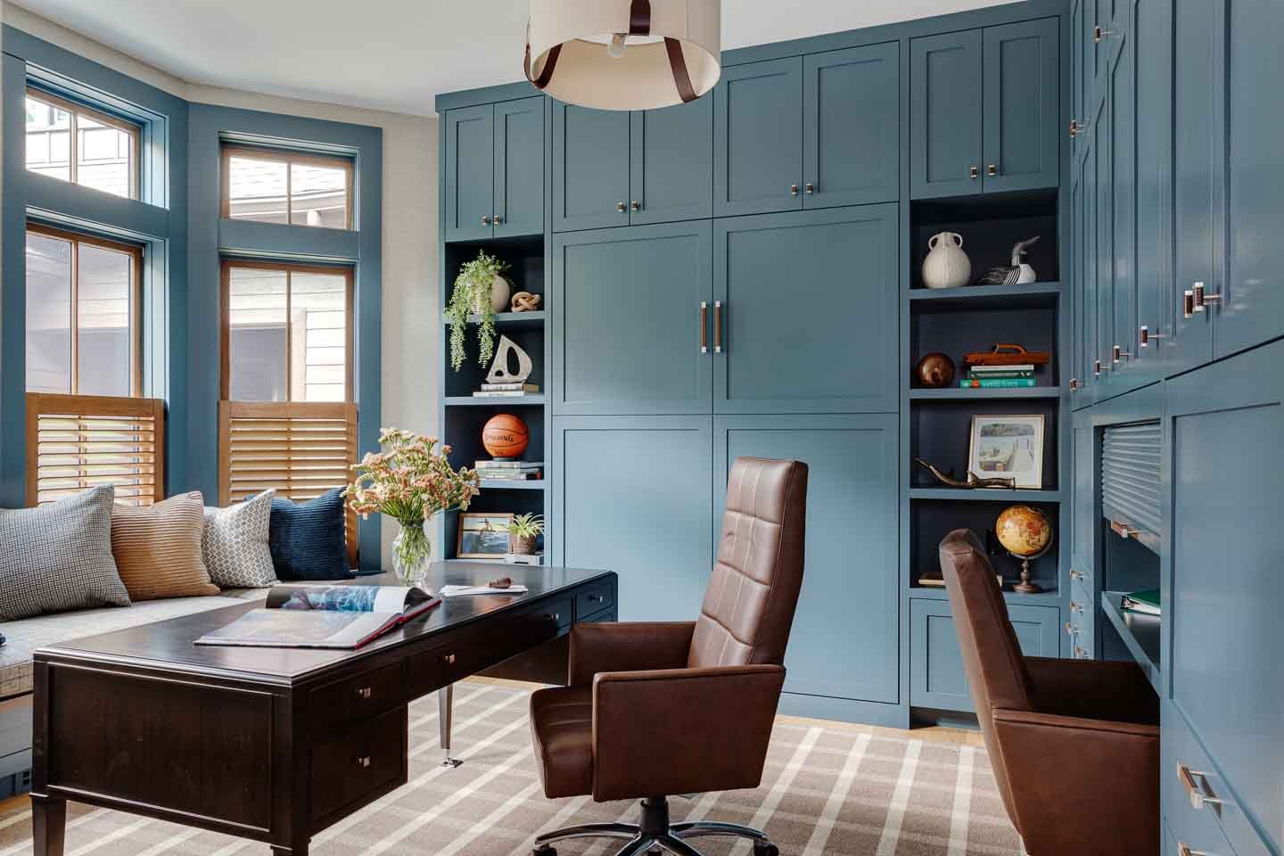 Office with brown leather desk chair, black desk, canvas light fixture, blue cabinets, built in window seat and plaid rug | Lindsay Lawrence Interiors