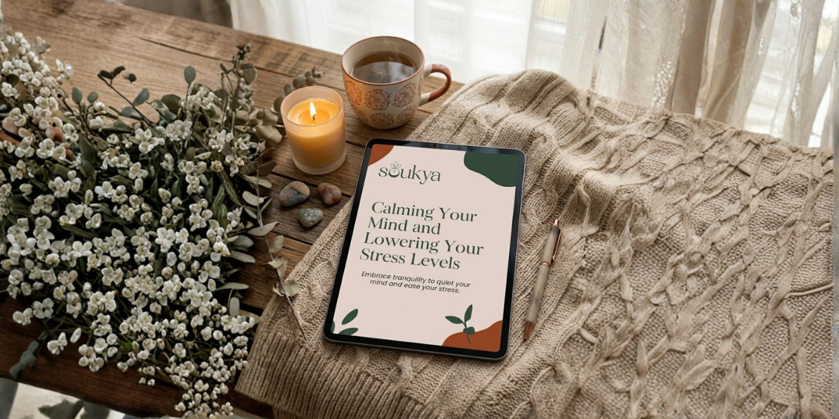 A cozy wooden table setup with a tablet displaying a calming mindfulness app, a lit candle, a cup of tea, a pen, a blanket, and a bunch of white flowers. Sunlight filters through curtains in the background.