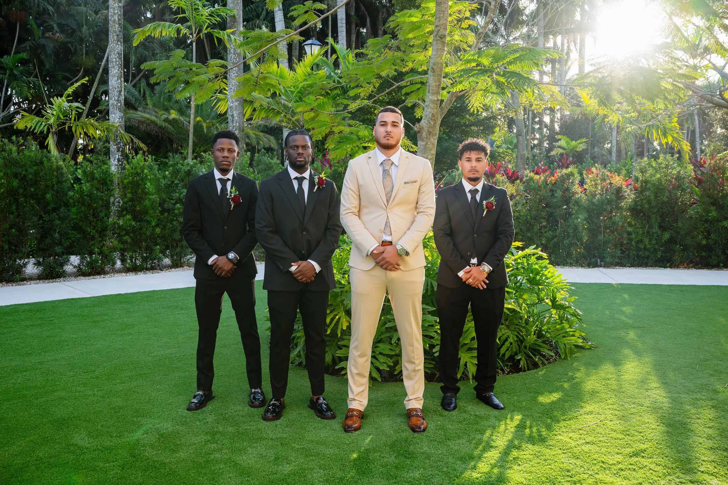 Four men dressed in formal suits posing outdoors on a lush green lawn with trees and bushes in the background.