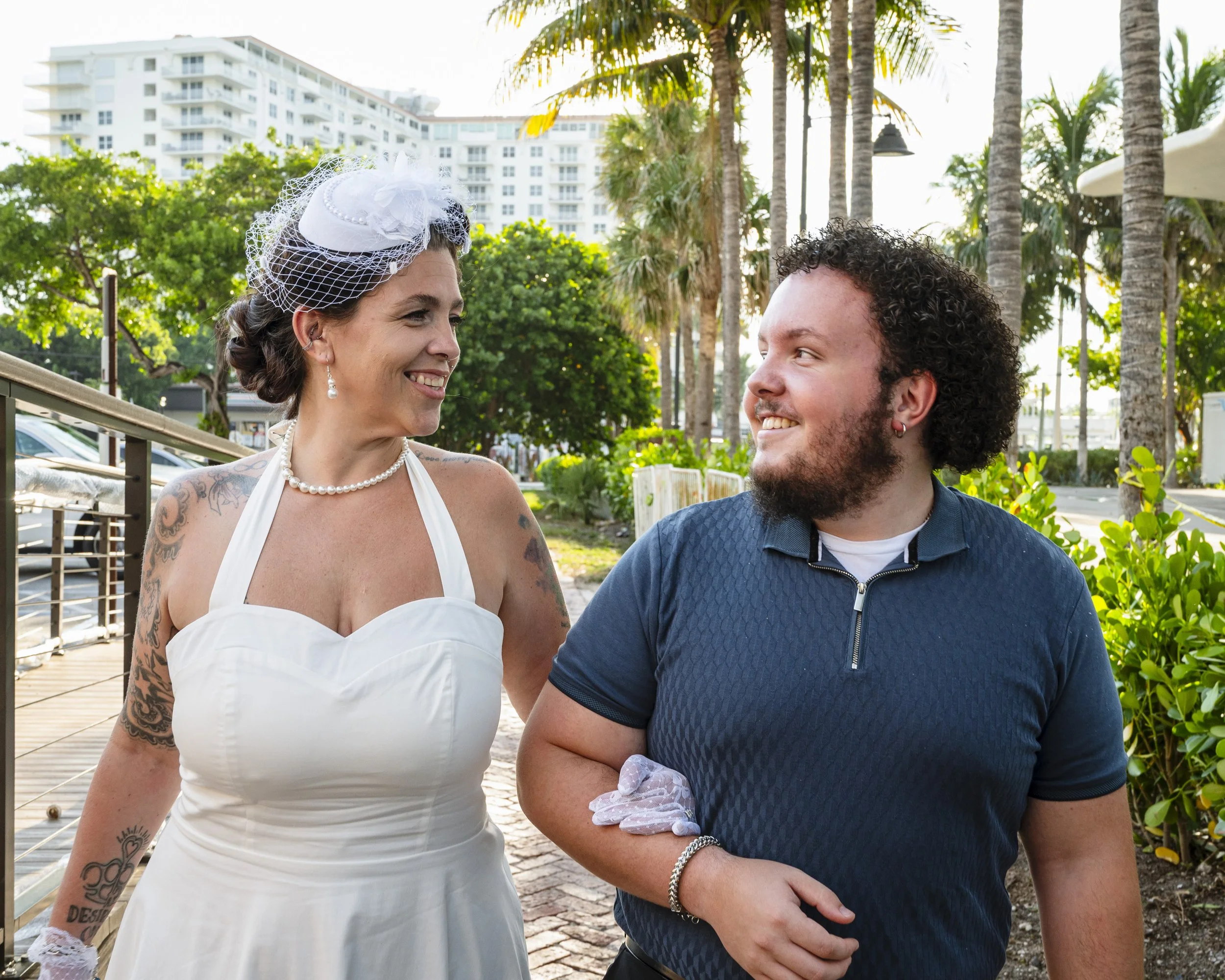 A woman in a white wedding dress with a veil and pearls, smiling at a man in a dark blue polo shirt holding her arm, outdoors with palm trees and high-rise buildings in the background.