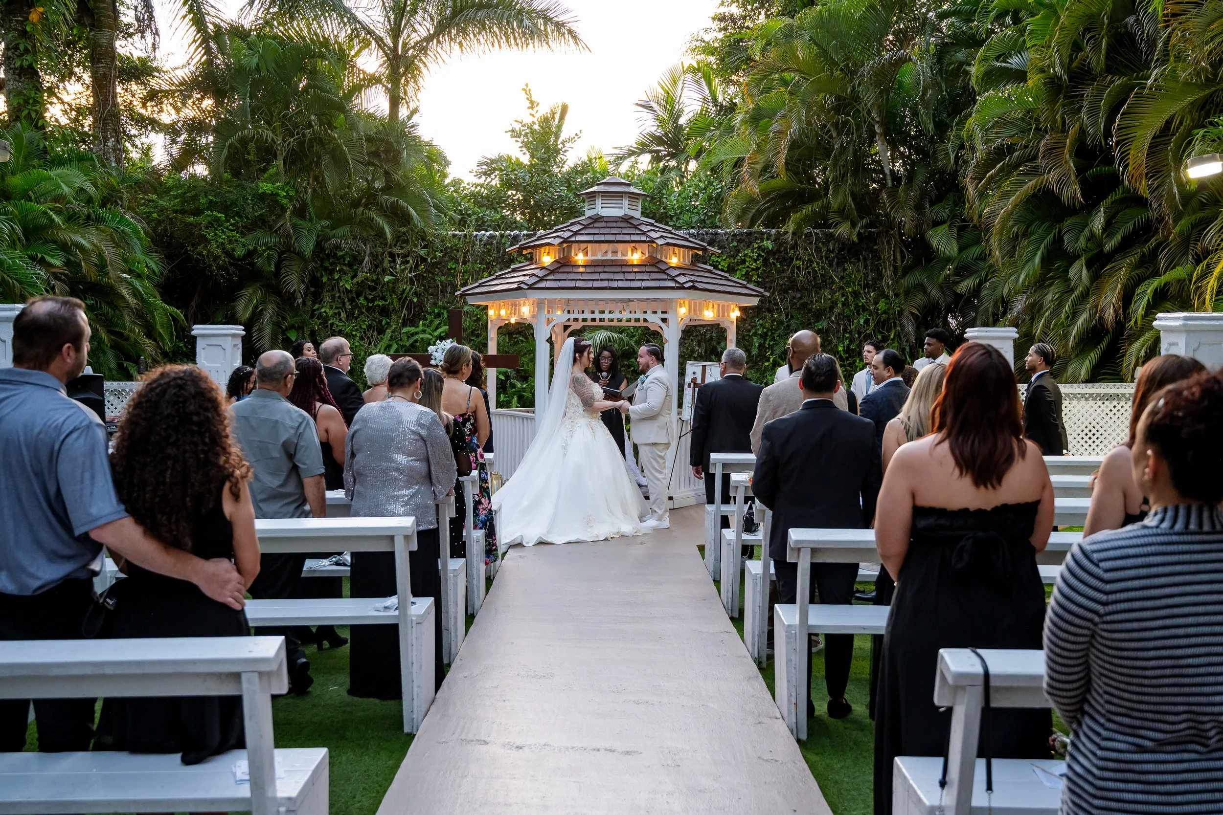 A wedding ceremony taking place outdoors in a lush garden with a wooden gazebo. The bride and groom are exchanging vows while guests are seated and standing on either side of the aisle. The scene is set during the evening with warm lighting and tall 