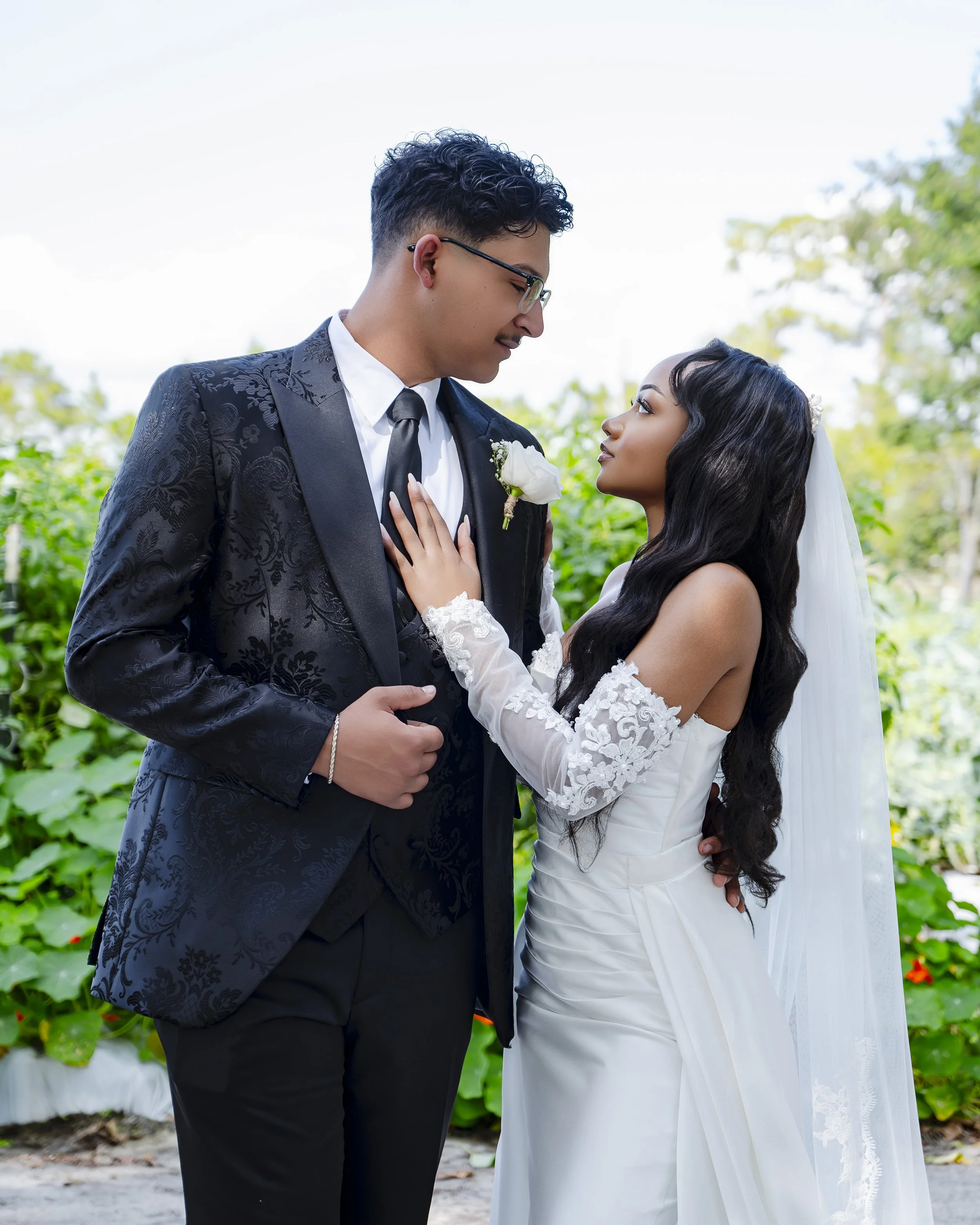 A bride and groom stand close together outside, gazing at each other. The groom wears glasses and a black tuxedo with a floral pattern, and the bride wears a white wedding dress with lace sleeves and a veil. The bride's hand is resting on the groom's