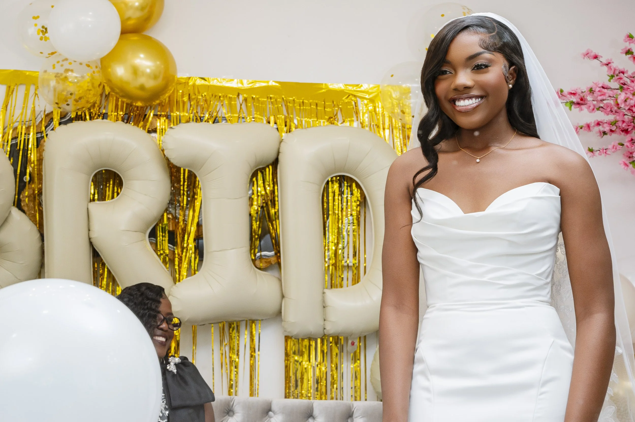 A woman in a white wedding dress is smiling at a celebration with gold and white balloons, large letter balloons spelling "PRIDE" in the background, and pink flowering trees.