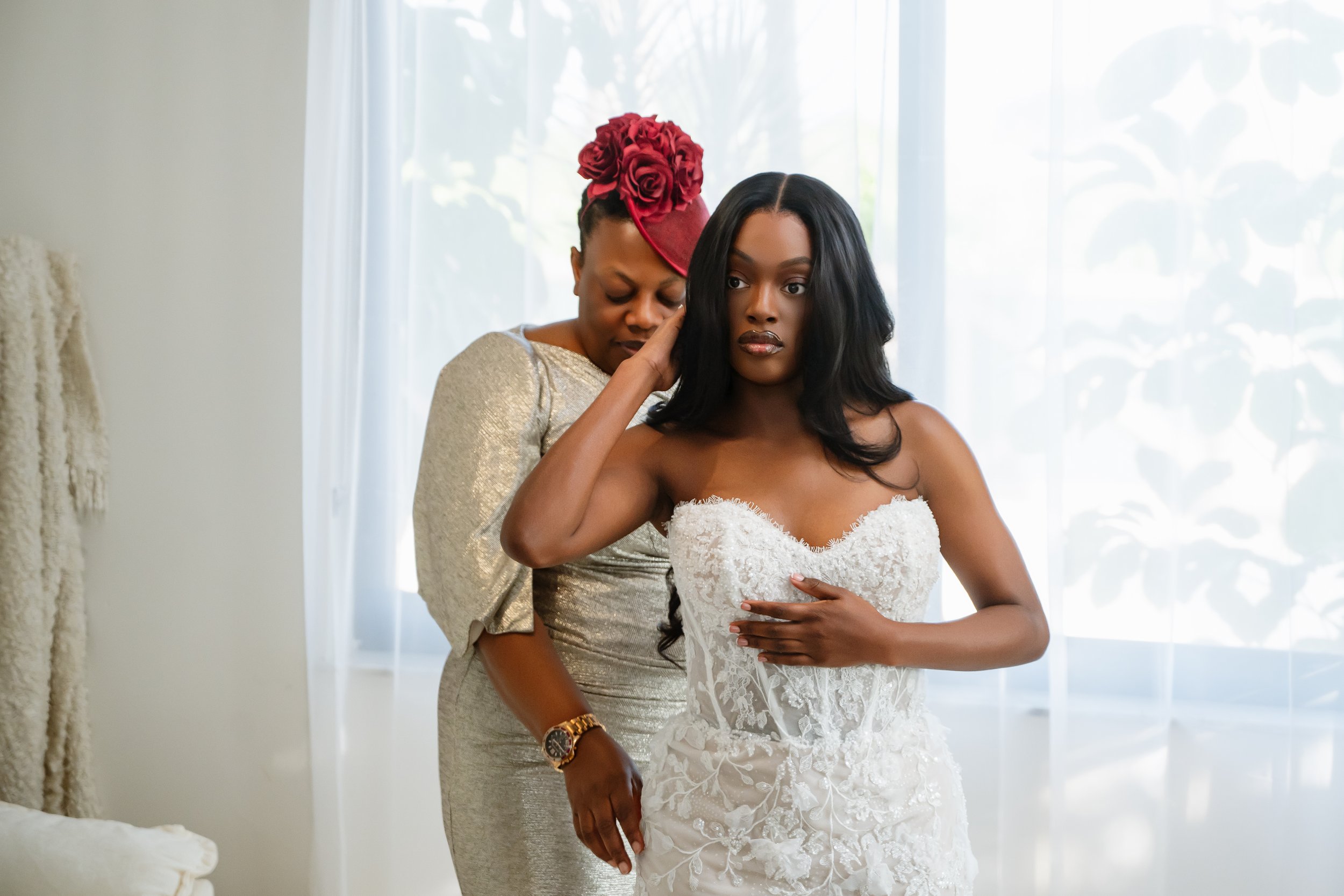 A bride in a white wedding gown with lace details standing in front of a window, with another woman behind her adjusting her dress. The woman behind is wearing a metallic dress and a floral headpiece.