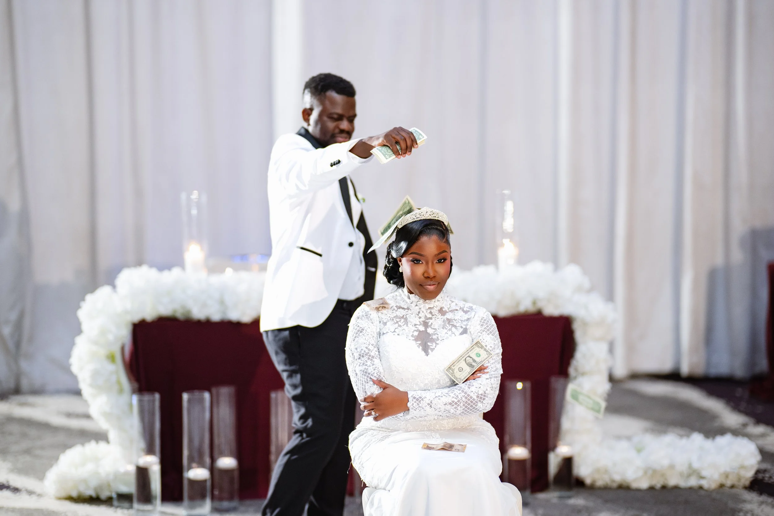 A couple performing a money dance at their wedding ceremony, with the groom standing and the bride sitting, both holding money. The bride is dressed in a white wedding gown with a tiara, and the groom is in a white tuxedo with black pants. The backgr