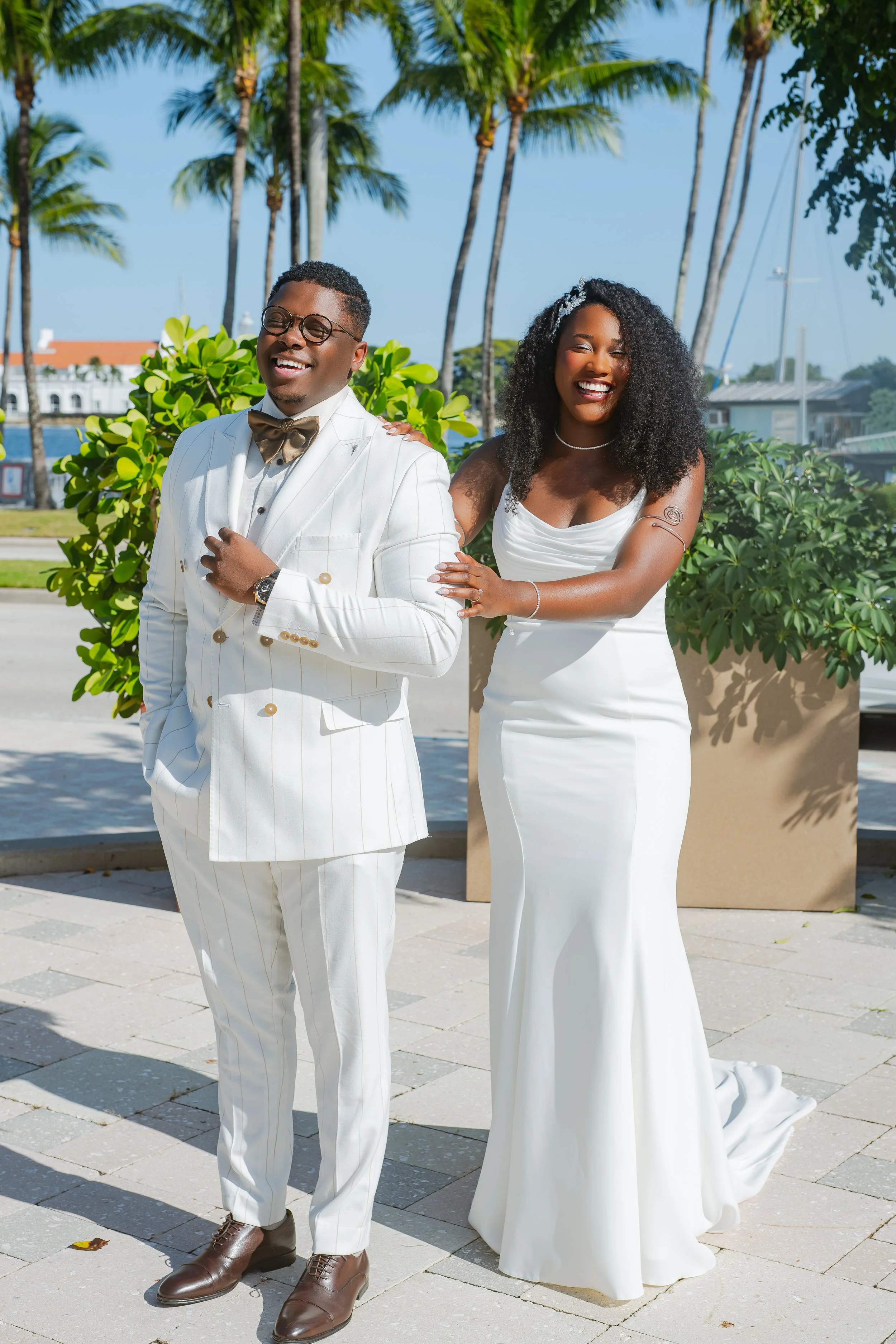 A newly married couple posing outdoors on a sunny day, with palm trees and boats in the background. The groom is wearing a white suit with a gold bow tie, and the bride is in a white wedding gown. Both are smiling, with the bride touching the groom's