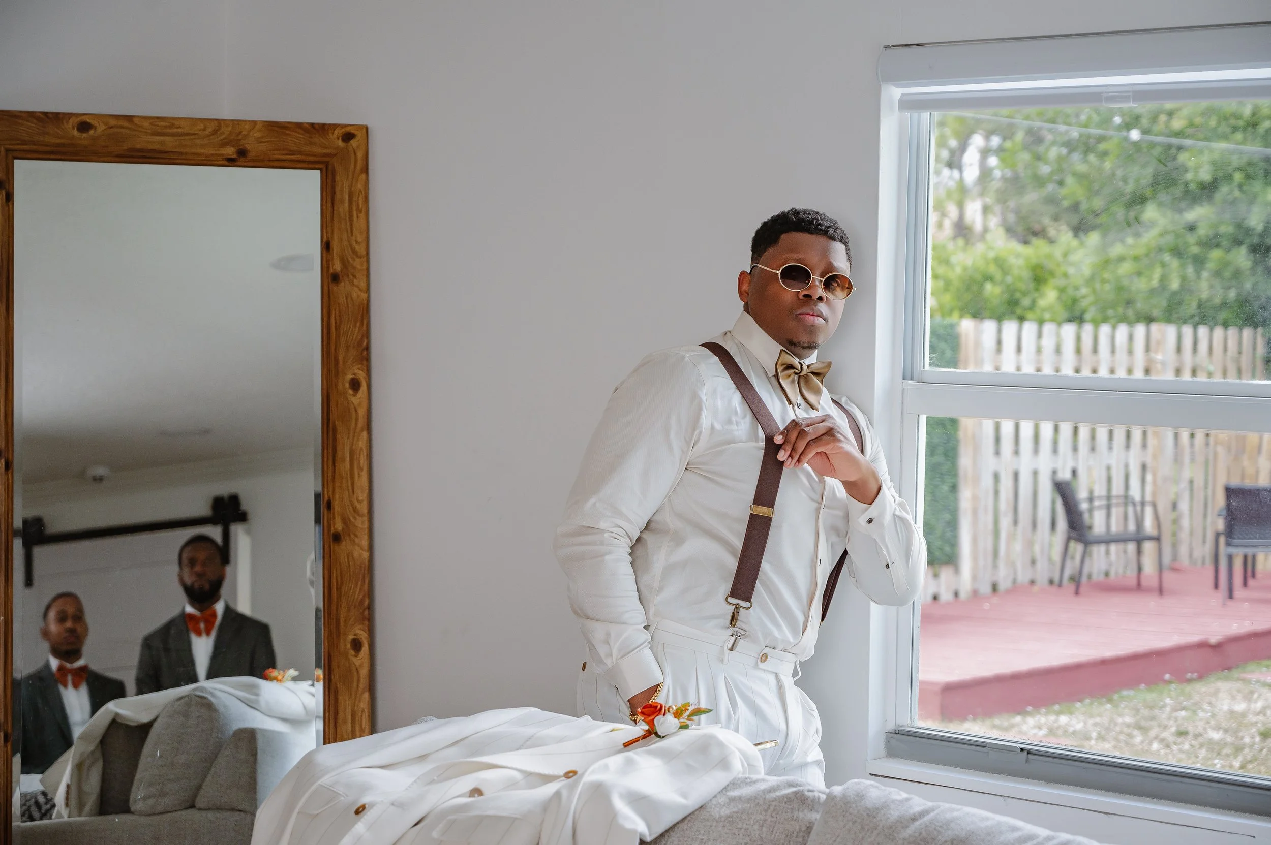 A man dressed in a white suit, suspenders, and a bowtie, wearing round sunglasses, standing near a window with a view of a backyard with chairs and a fence. Two men dressed in tuxedos with orange bowties, visible in the mirror reflection.