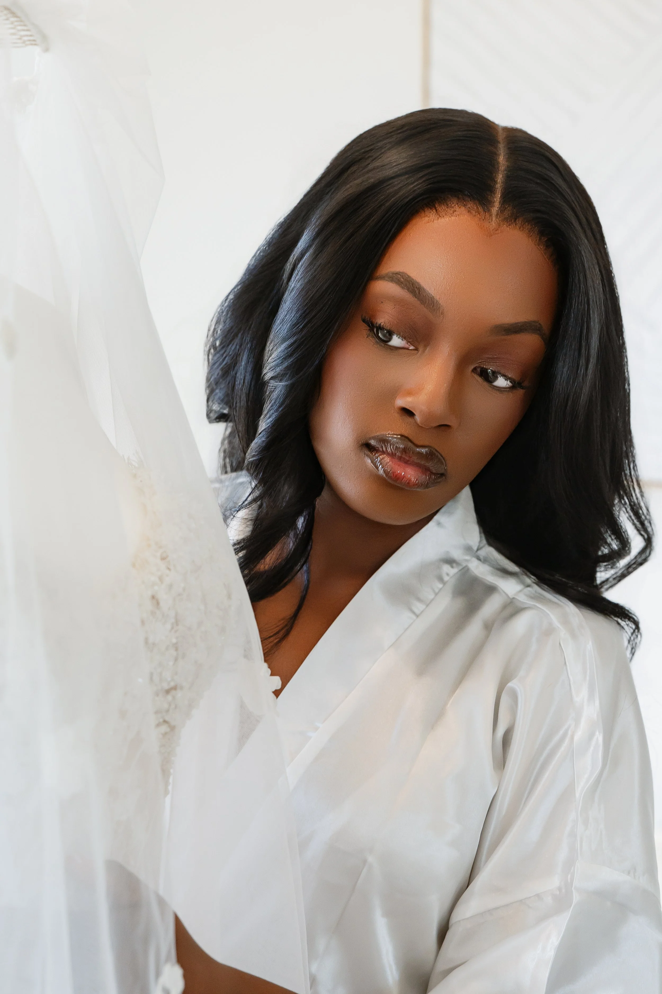 A woman with dark skin and black hair styled in loose waves, wearing a white satin robe, looking to the side with a neutral expression.