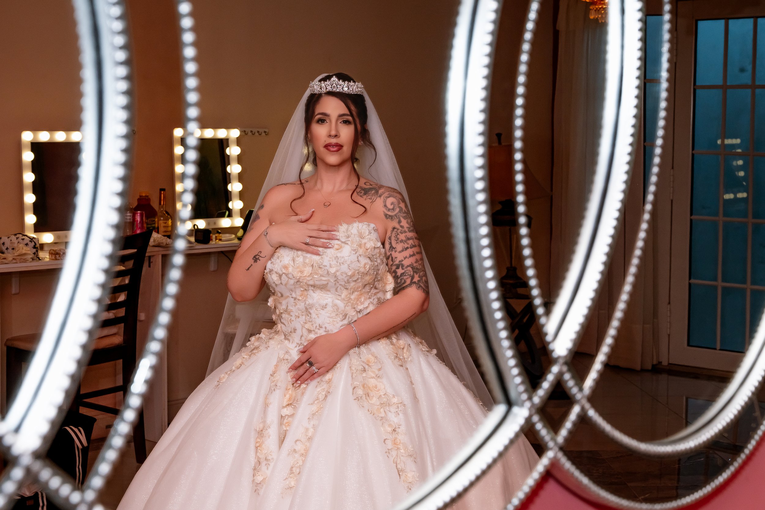A woman in a wedding dress and veil, wearing a tiara, standing in front of a mirror, with her hand on her chest, in a room with makeup mirrors and vanity items.
