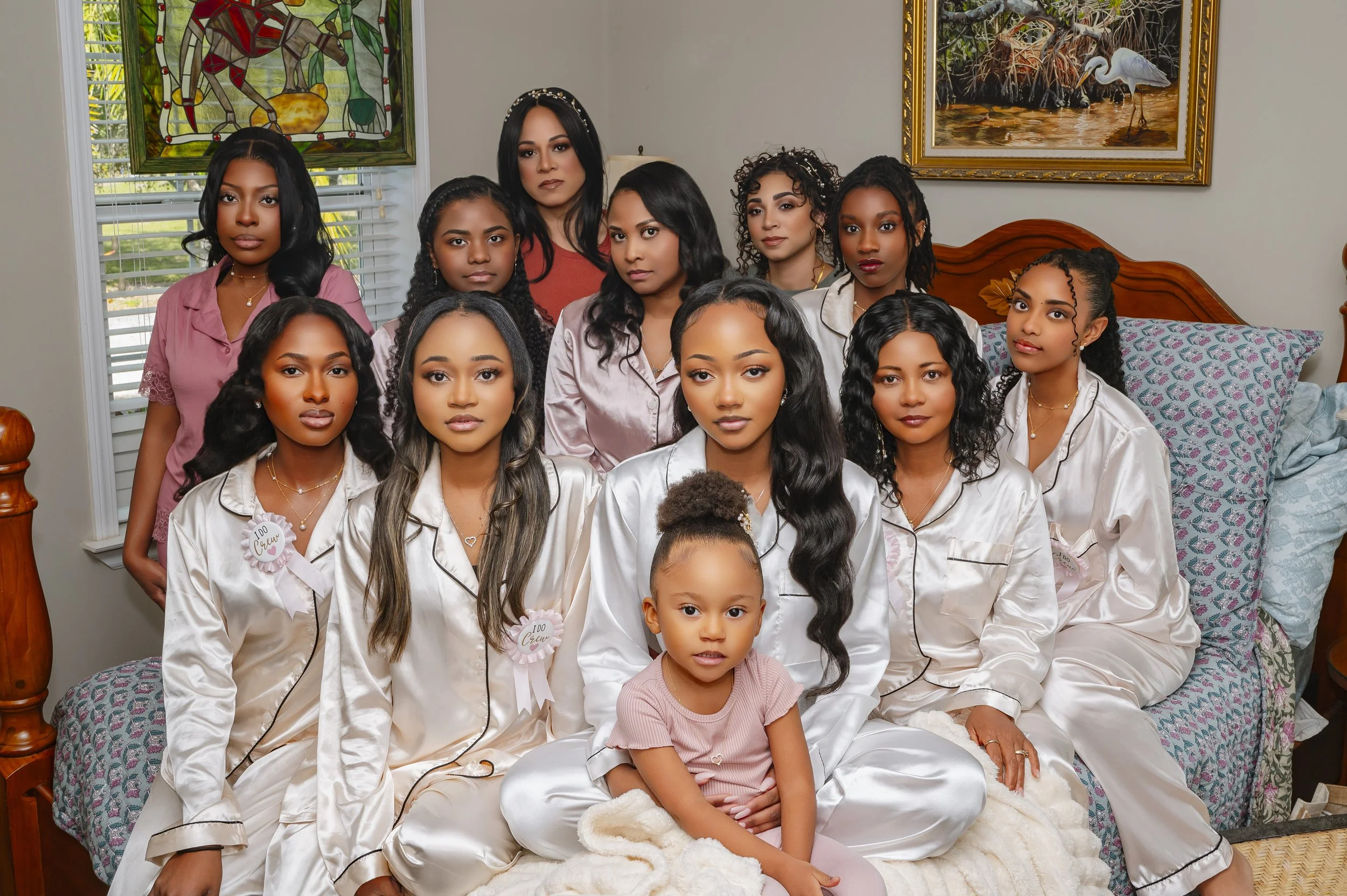 A group of 12 women and a young girl in pajamas, sitting and standing on a bed in a bedroom. The women are wearing satin pajamas in white, pink, and beige, with some having name tags or ribbons. The young girl in the front is wearing a pink shirt and