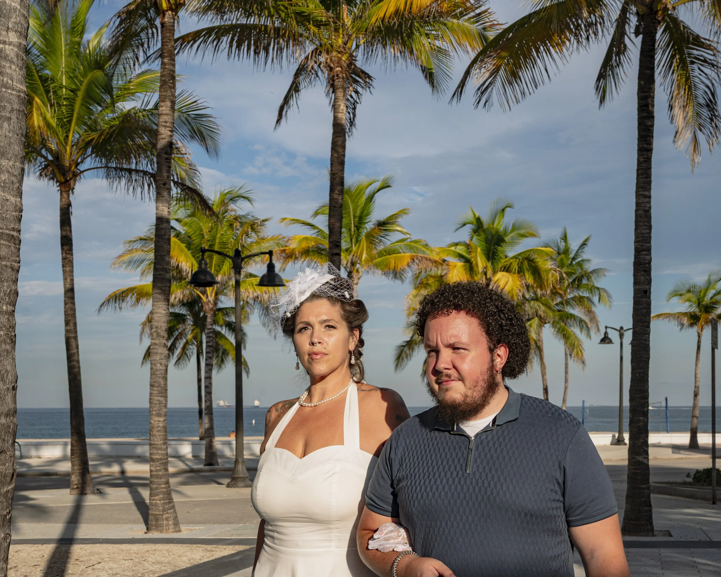 A woman in a white dress and a man with curly hair and beard standing outdoors in front of palm trees on a sunny day, near a beach.