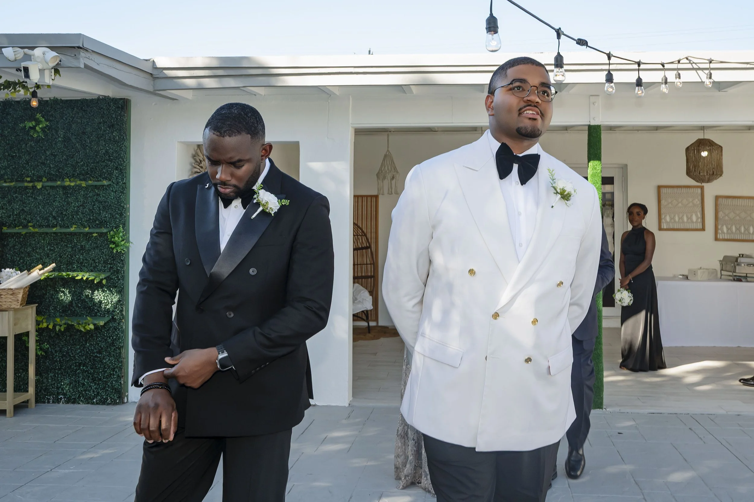 Two men dressed in formal suits at a wedding, one in a black tuxedo looking down, and the other in a white tuxedo with a bow tie looking forward, with a woman in a black dress holding a small bouquet in the background under string lights.
