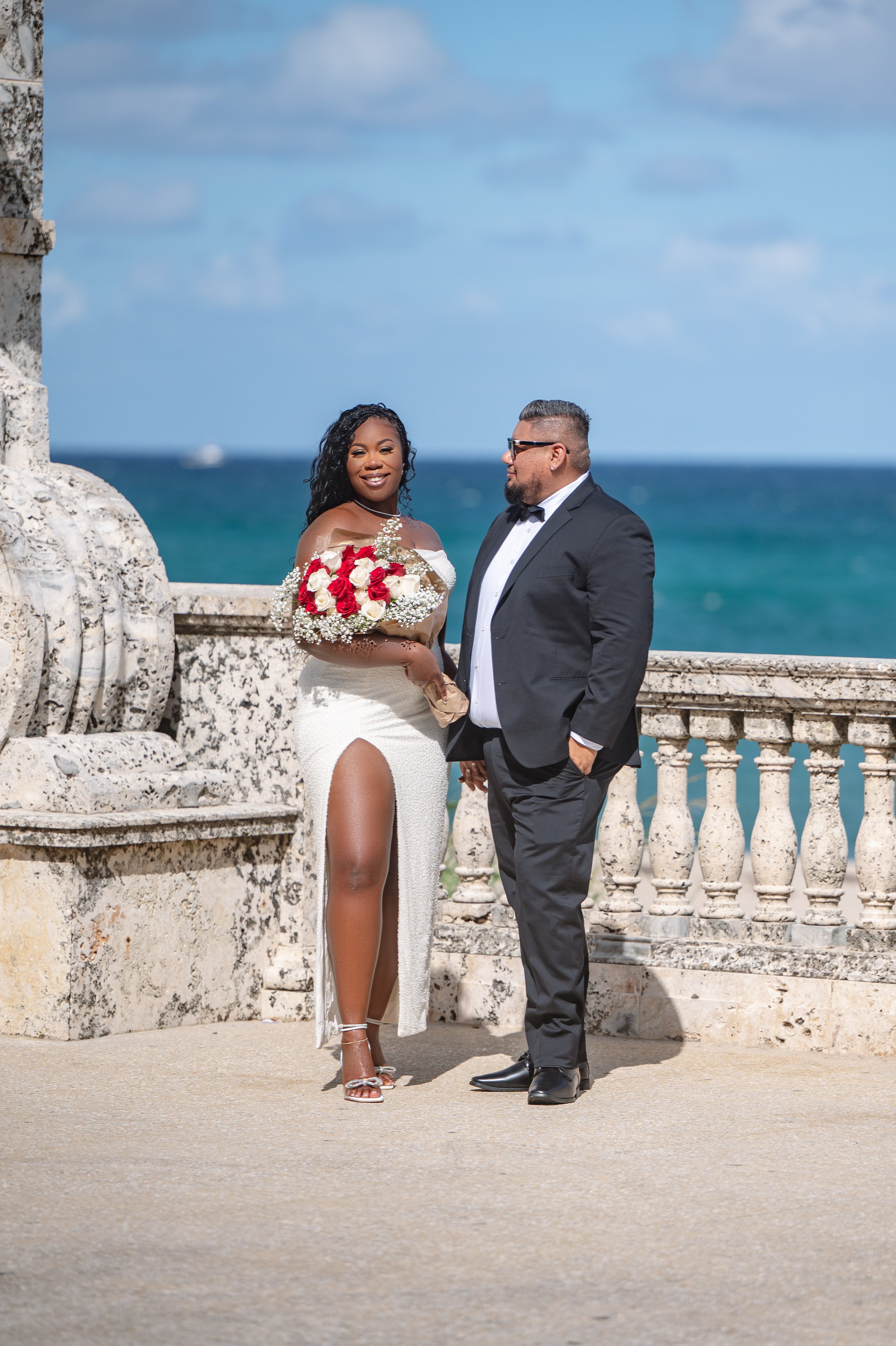 A bride and groom standing on a terrace near the ocean, with a stone railing and blue sky in the background. The bride is holding a bouquet and wearing a white dress with a high slit, while the groom is dressed in a black tuxedo.