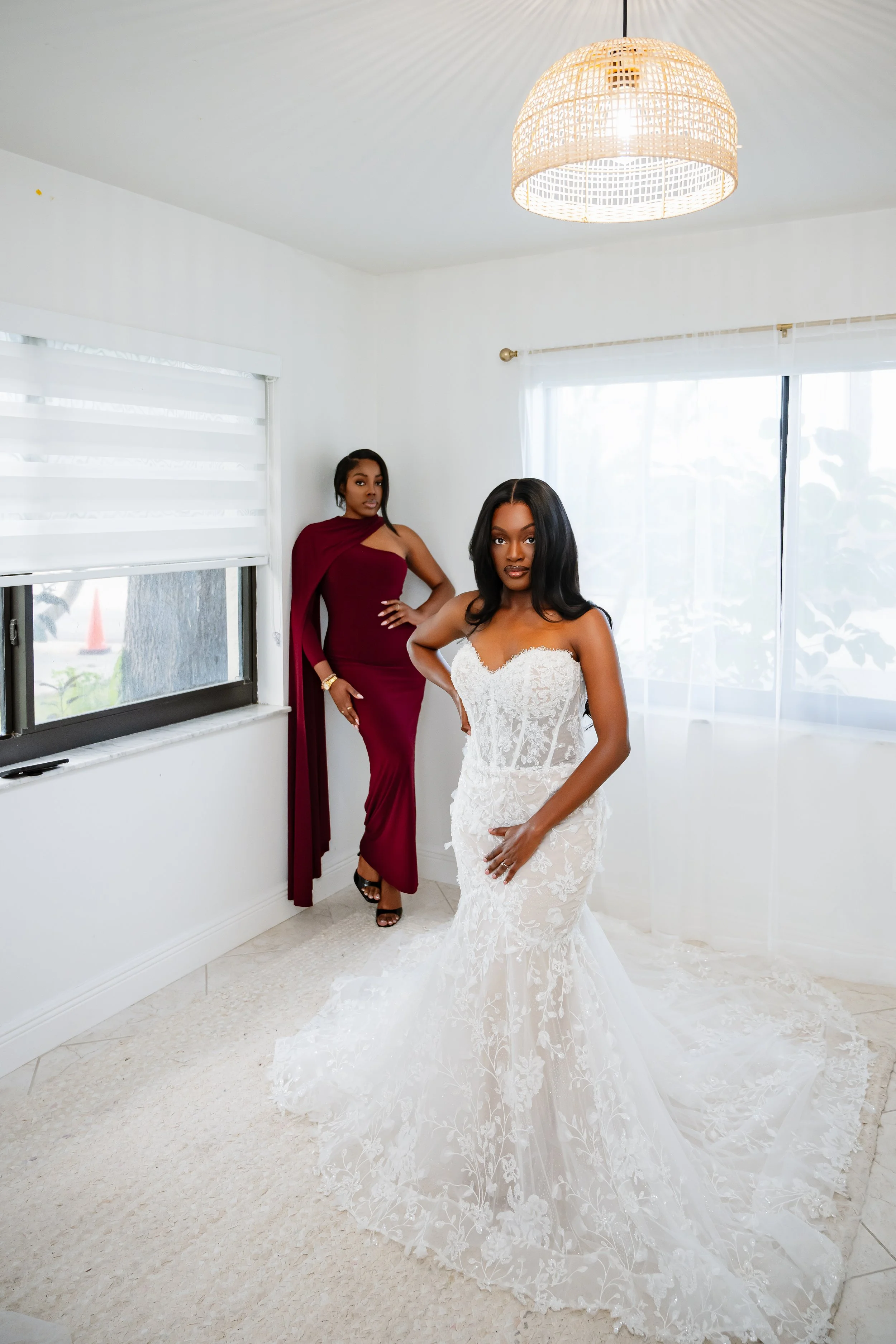 Two women, one in a white wedding dress and the other in a maroon gown, standing in a bright room with large windows and white curtains.