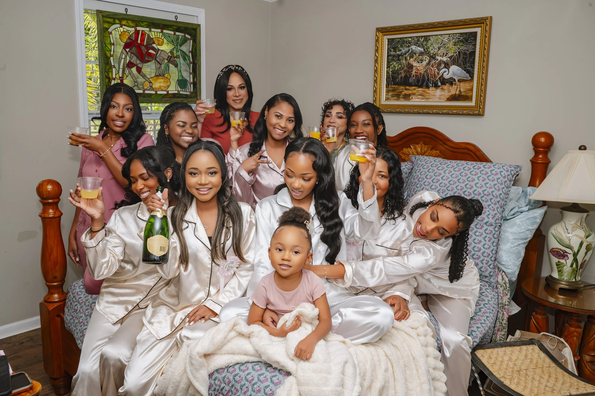 Group of women and a young girl celebrating in pajamas, some holding glasses with drinks, inside a bedroom with a bed, pillows, paintings, and a lamp.