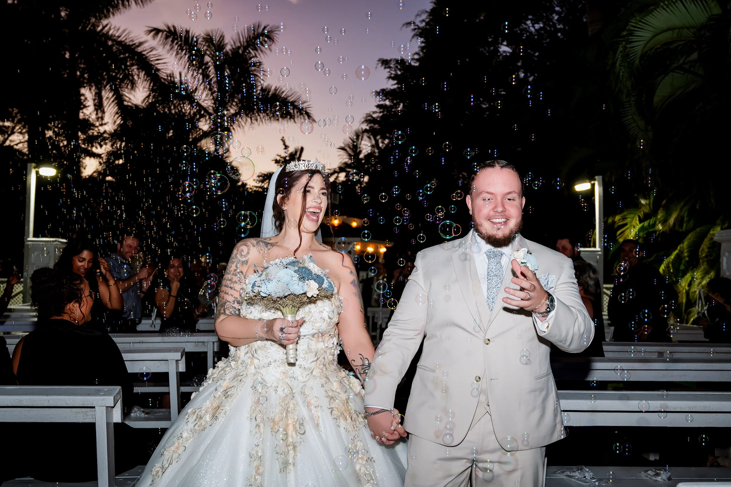 A newlywed couple holding hands and smiling during their wedding celebration, surrounded by bubbles and guests in an outdoor venue at dusk.