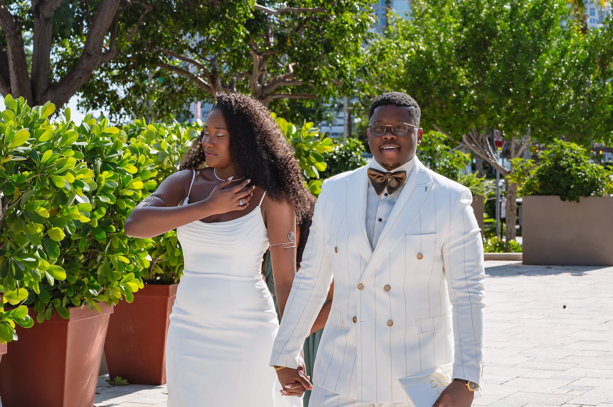 A man and woman dressed in formal attire holding hands outdoors during the daytime. The woman is wearing a white dress and has curly hair, while the man is dressed in a white suit with gold accents, a bow tie, and glasses. The woman appears to be tou