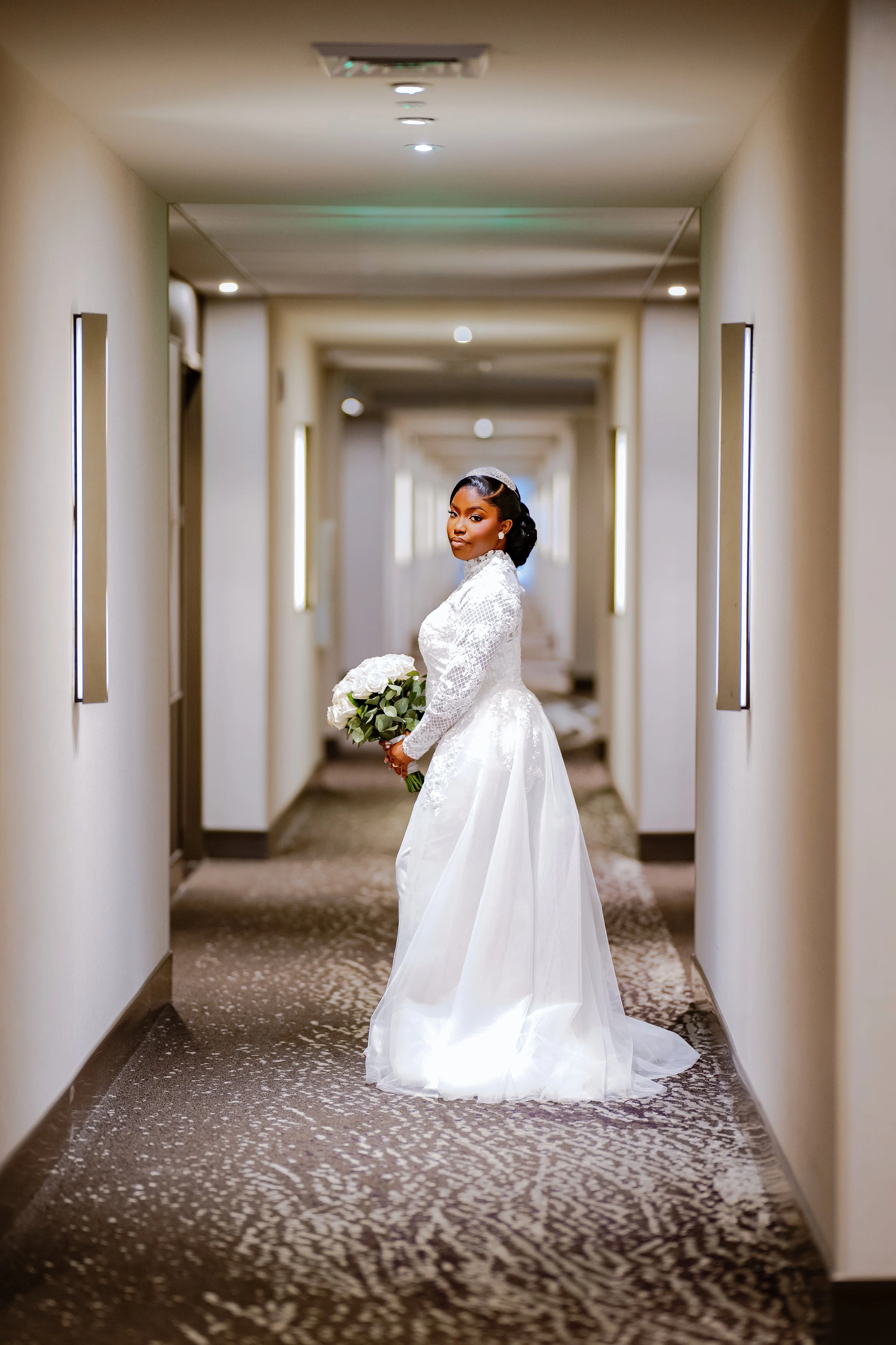 A bride in a white wedding gown standing in a hallway, holding a bouquet of white roses, with her head slightly turned to the side.