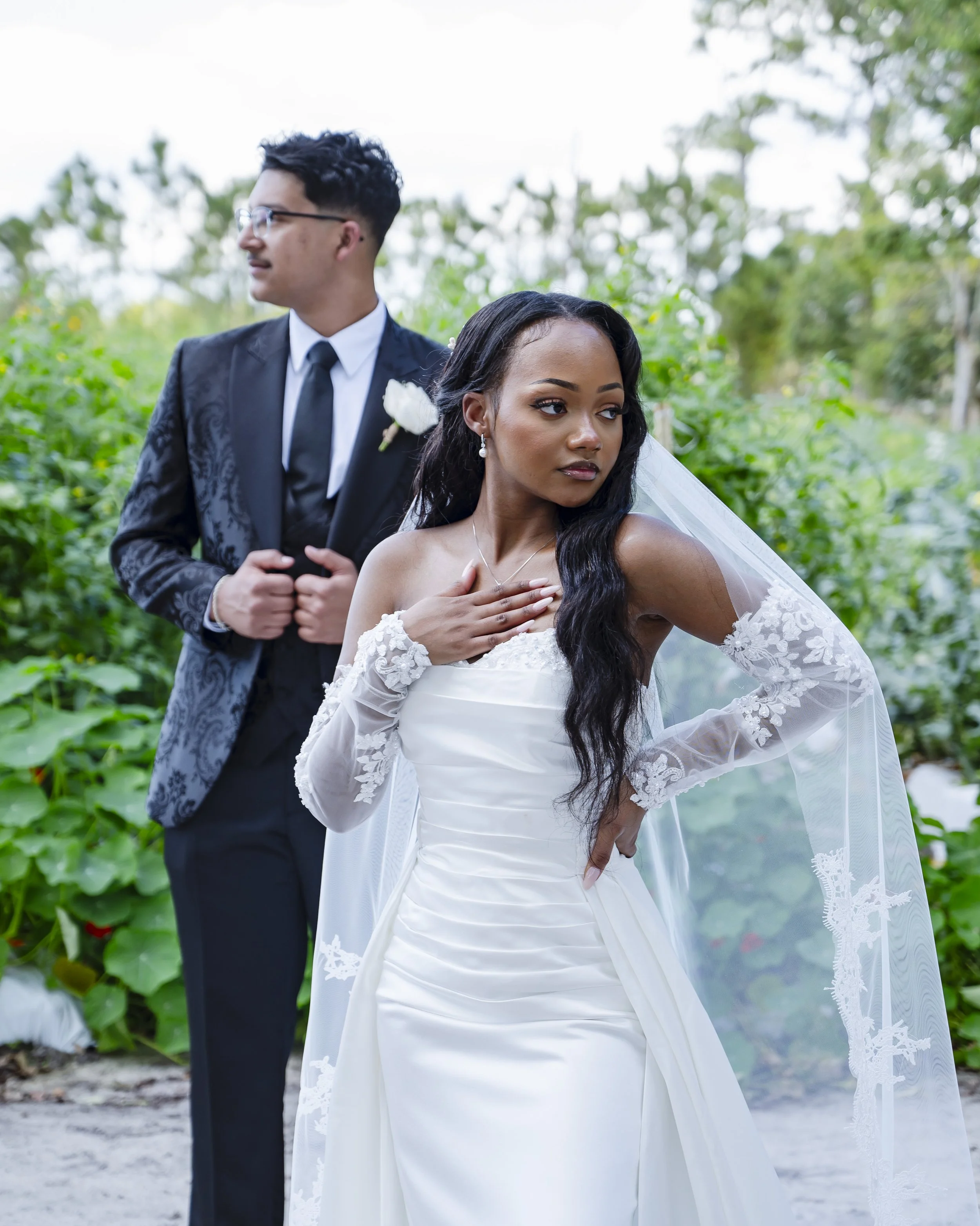 A bride in a white wedding gown and a groom in a black tuxedo pose outdoors in a lush green setting.
