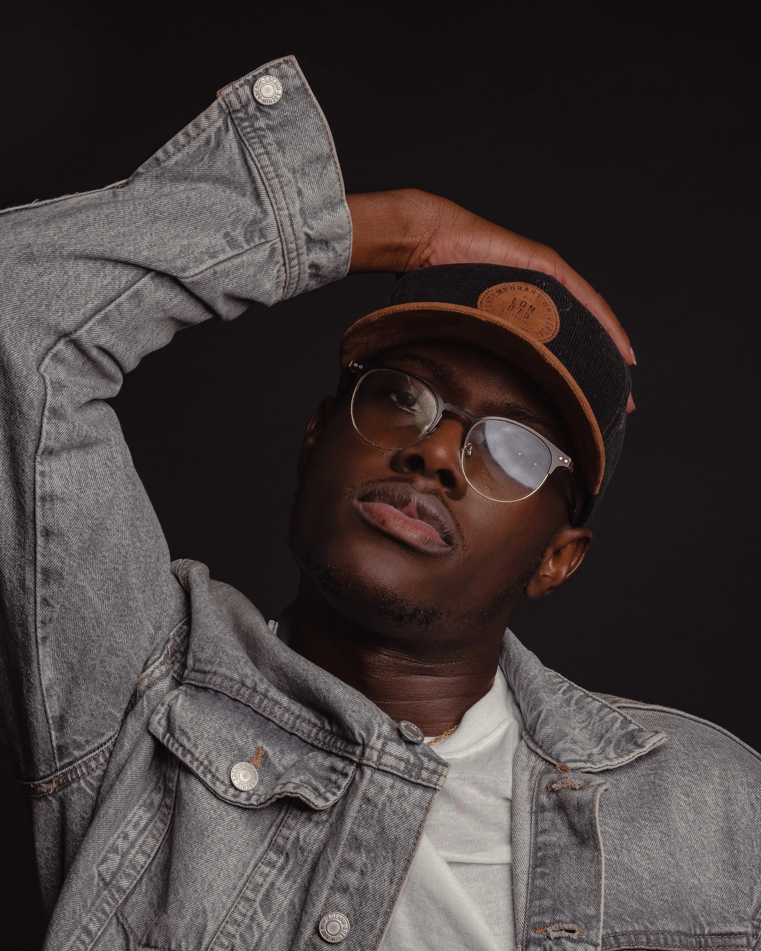 Portrait of a man wearing glasses, a denim jacket, a white t-shirt, and a black cap with a brown leather patch, against a black background.