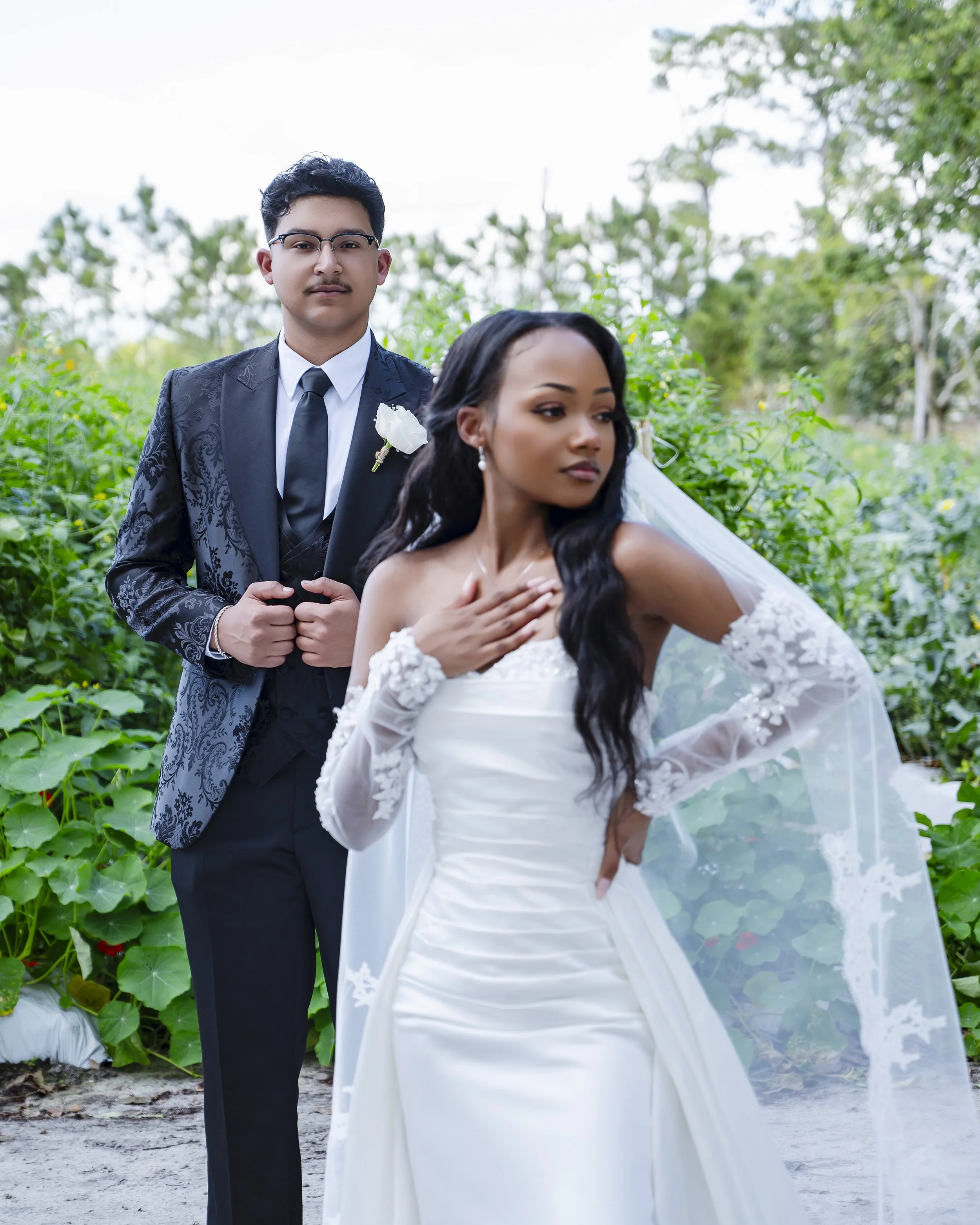 A bride and groom posing outdoors, with the bride in a white wedding dress with lace details and a long veil, the groom in a black suit with a white rose boutonniere, surrounded by greenery.