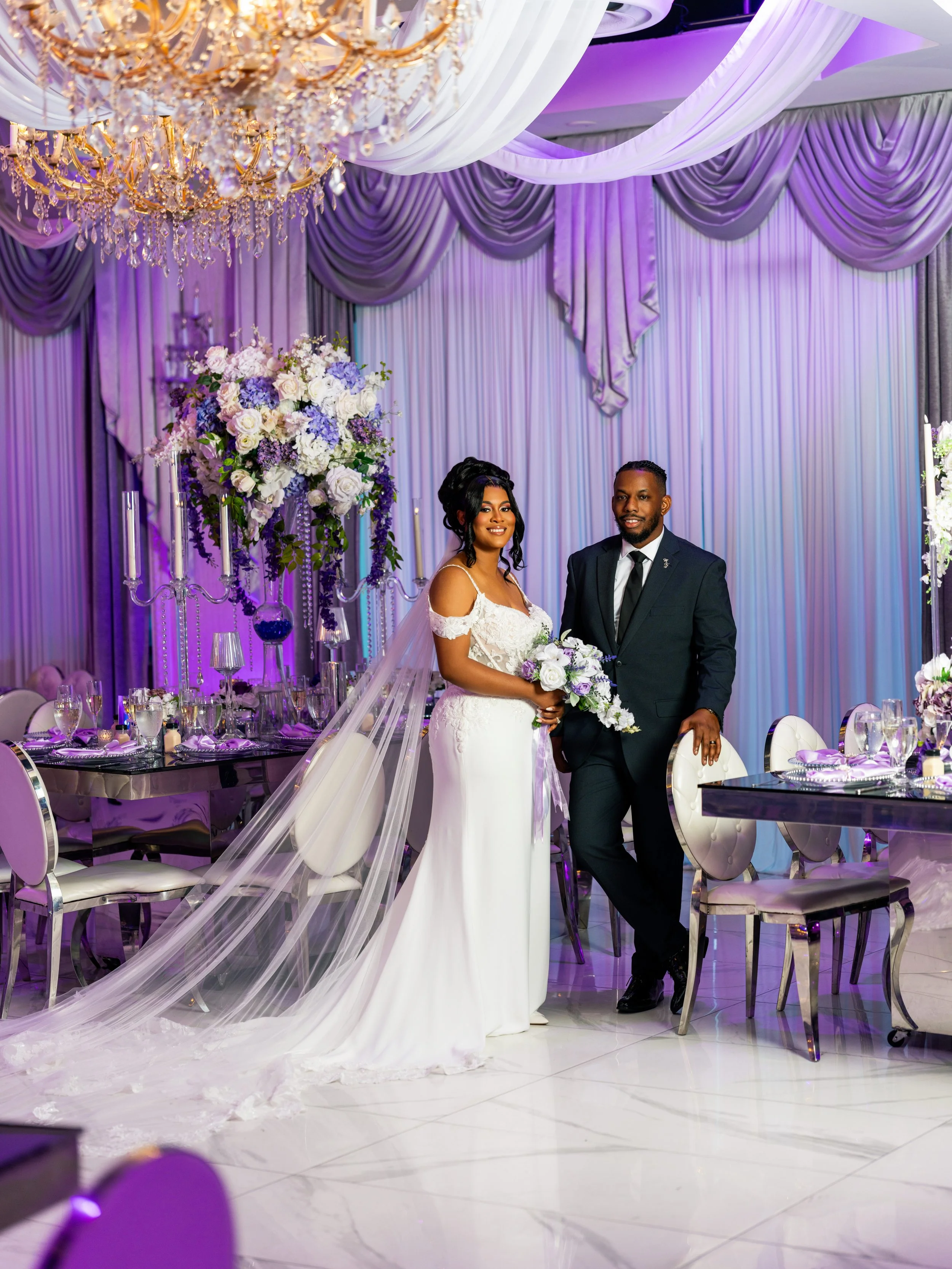 Elegantly decorated wedding reception with a bride and groom standing together, surrounded by purple and white draped curtains, a large floral arrangement, and a grand crystal chandelier.