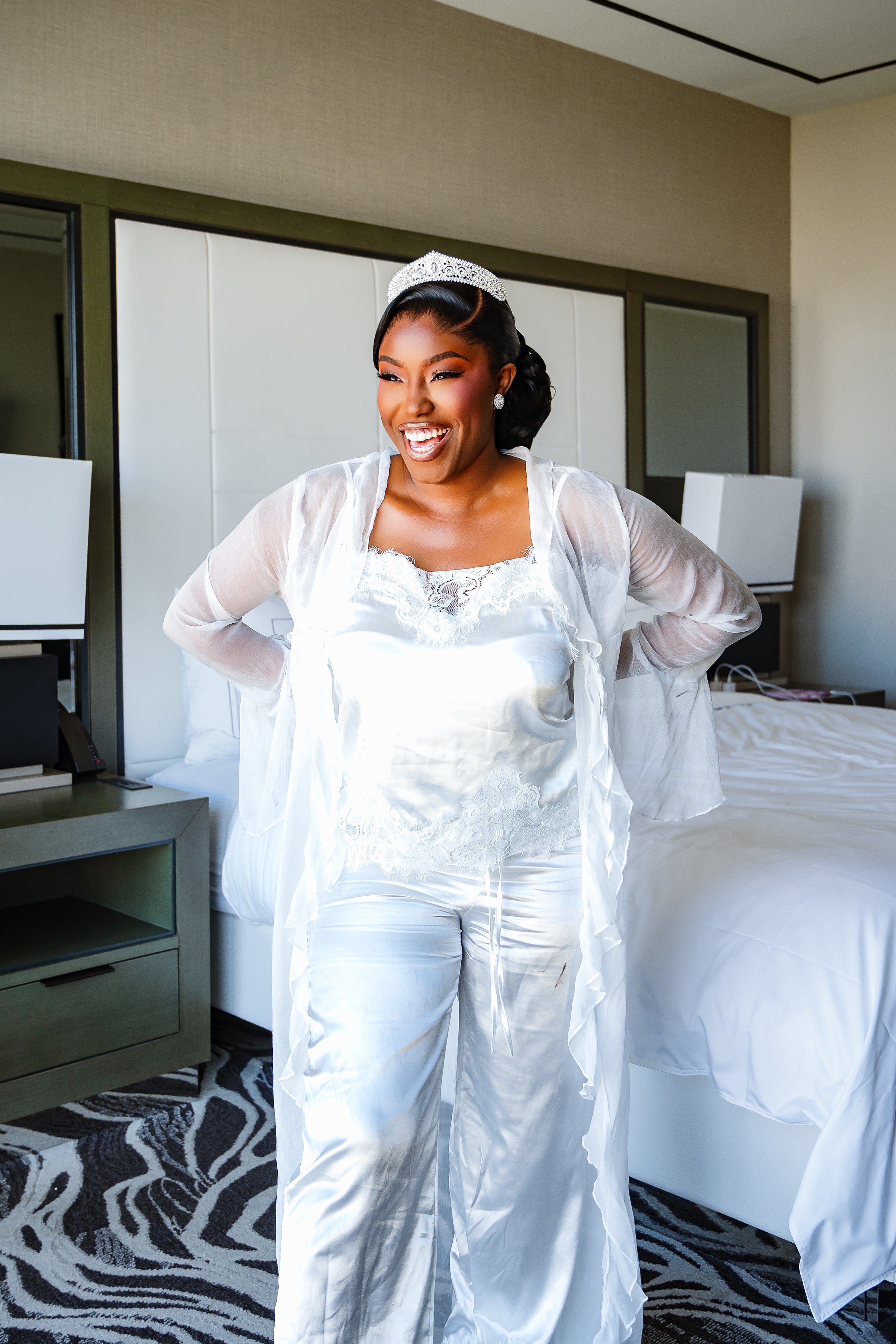 A woman wearing a white lacy top, satin pajamas, a tiara, and jewelry, smiling and standing in a hotel room near the bed.